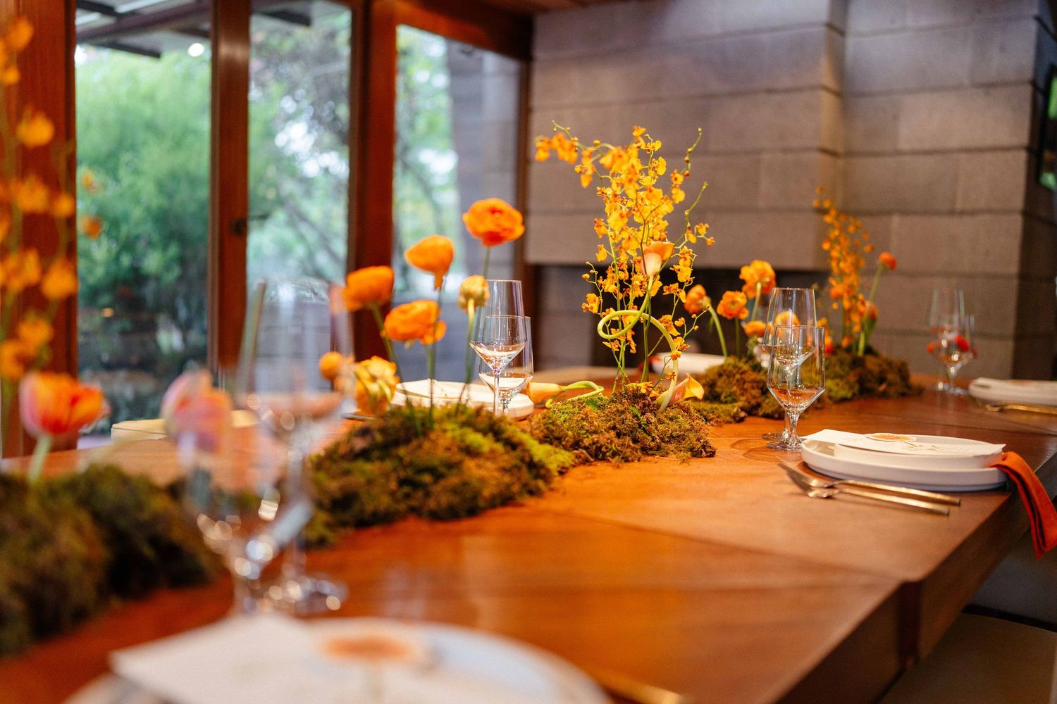 Close-up of wedding reception tablescape with orange ranunculus, moss runner, and glassware on a warm wood table.