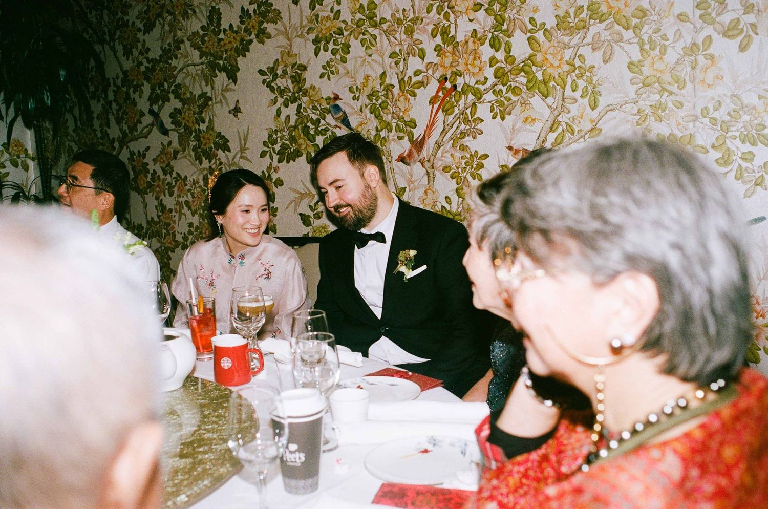 Bride and groom laughing at a floral wallpapered wedding reception dinner table with guests.