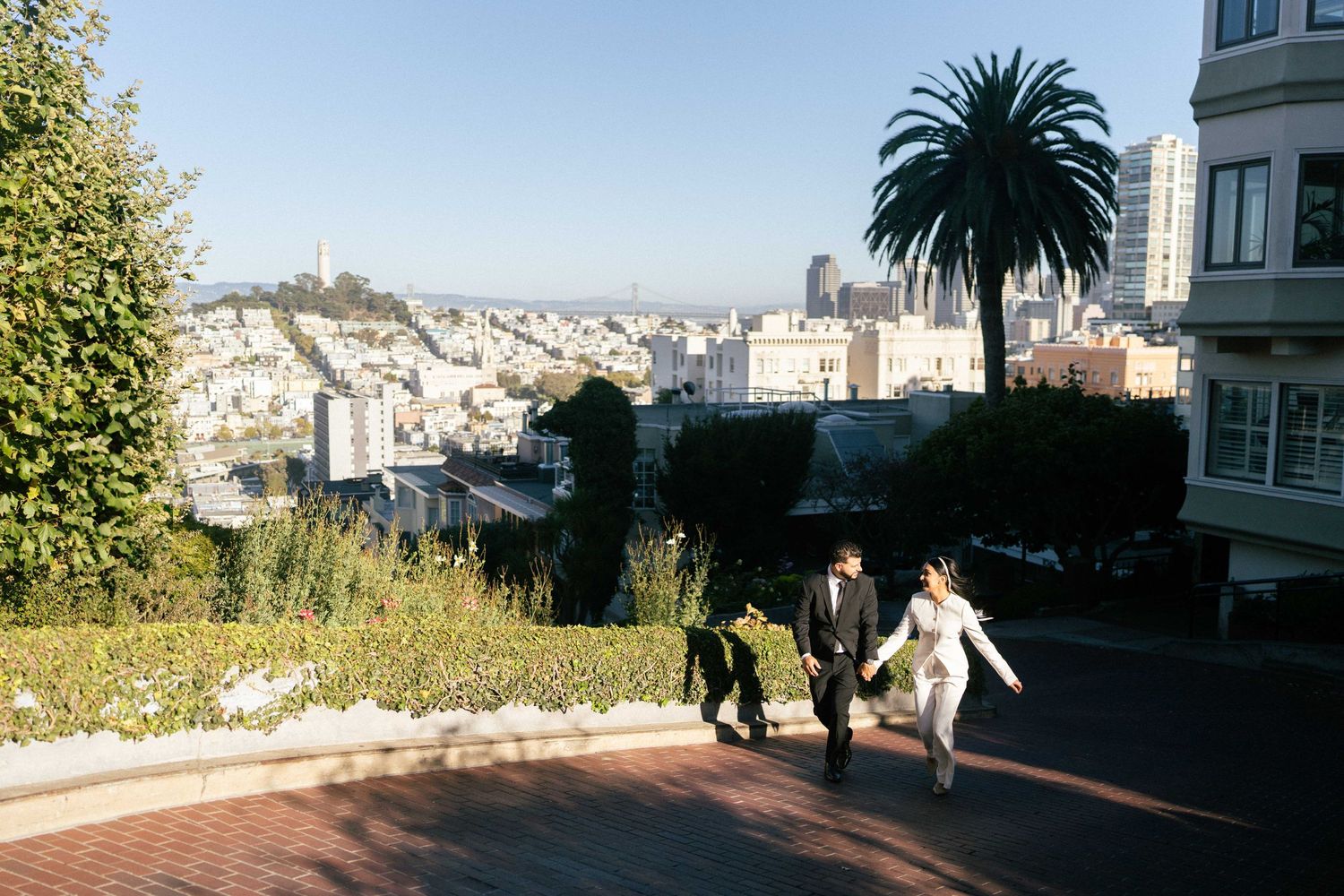 Wedding couple ascending steep streets of San Francisco with city panorama and palm tree in background.