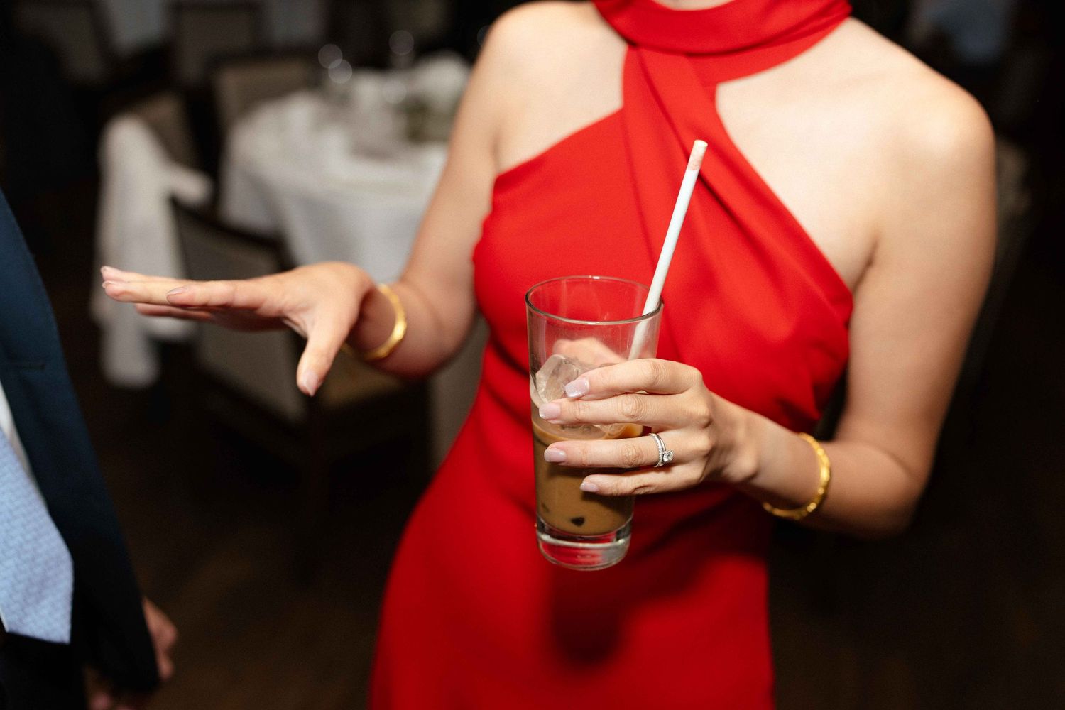 Someone in a red halter dress holds a beverage while gesturing at an indoor event.