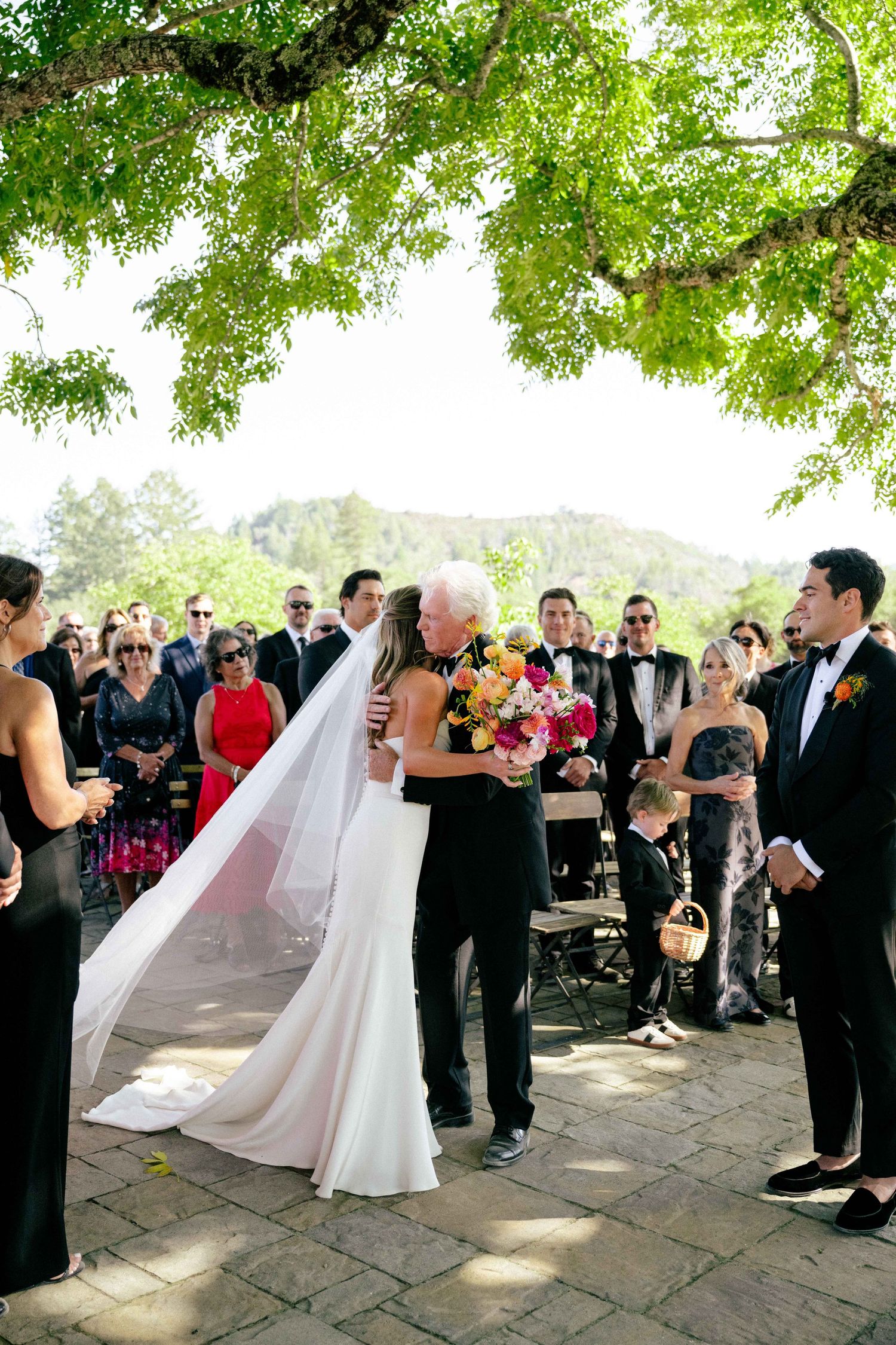 Father of the bride sharing an emotional embrace at the wedding ceremony under a large tree.