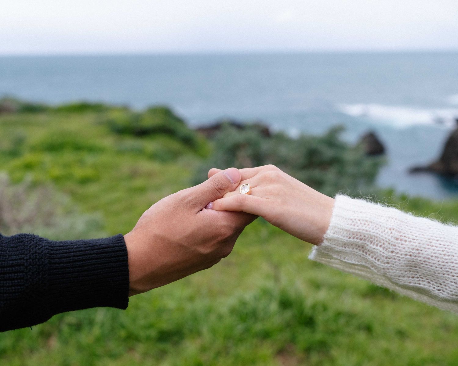 Couple holding hands with engagement ring near ocean cliffs and green grass backdrop.