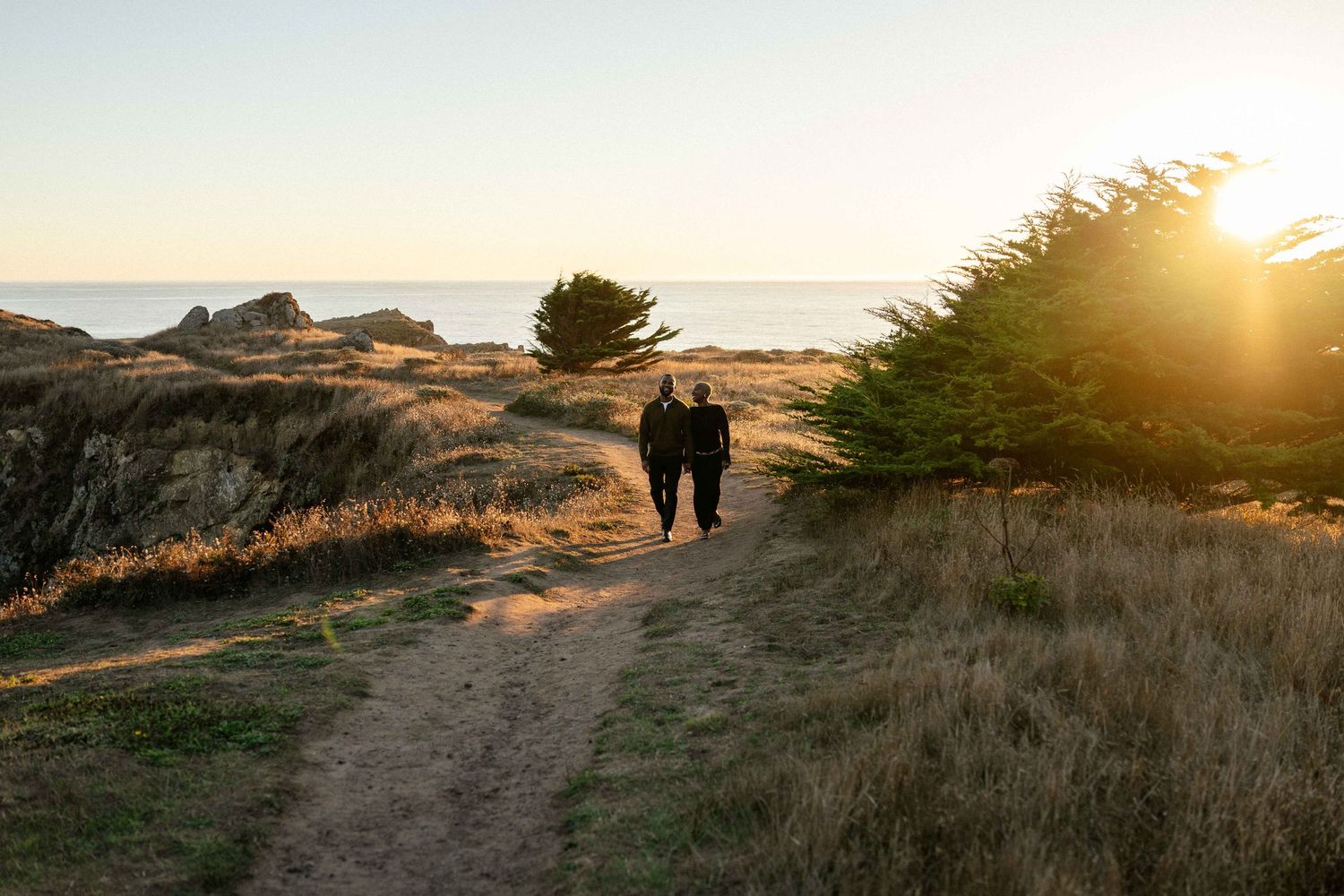 Two people walk hand in hand on a coastal trail at golden sunset, surrounded by grassy dunes and windswept trees.