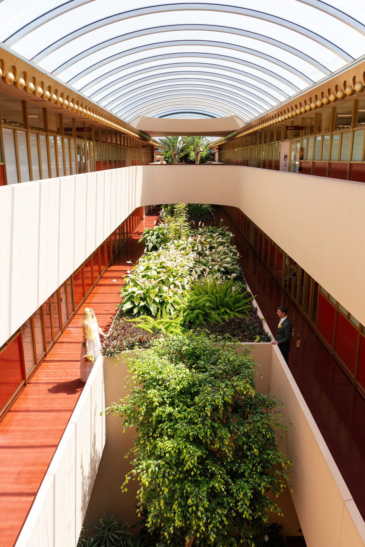 Modern atrium with lush green plants, red walkways, and a curved glass ceiling in a contemporary building interior.