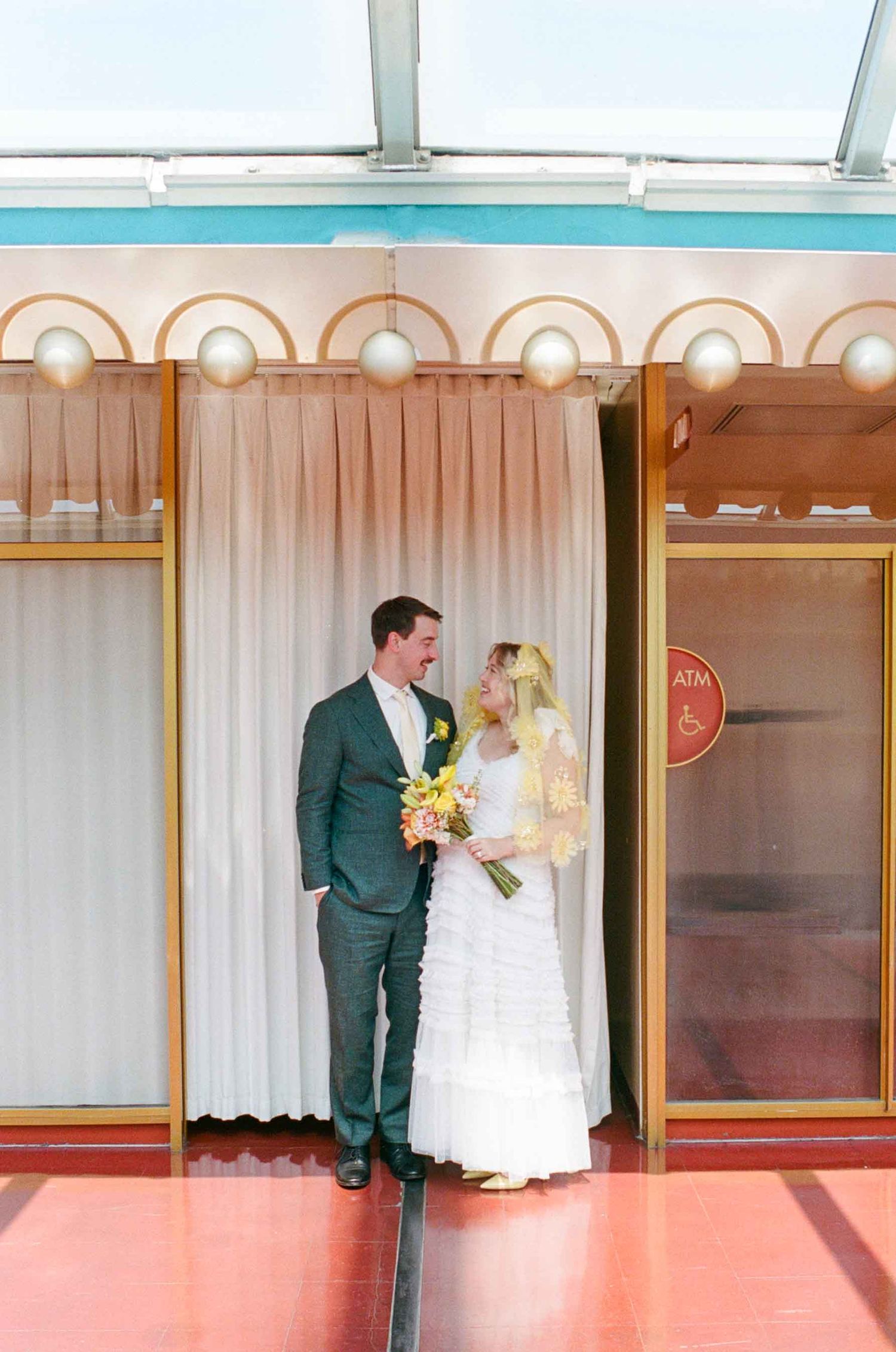 Bride and groom pose together in a colorful retro venue, bride holding a vibrant bouquet on their wedding day.