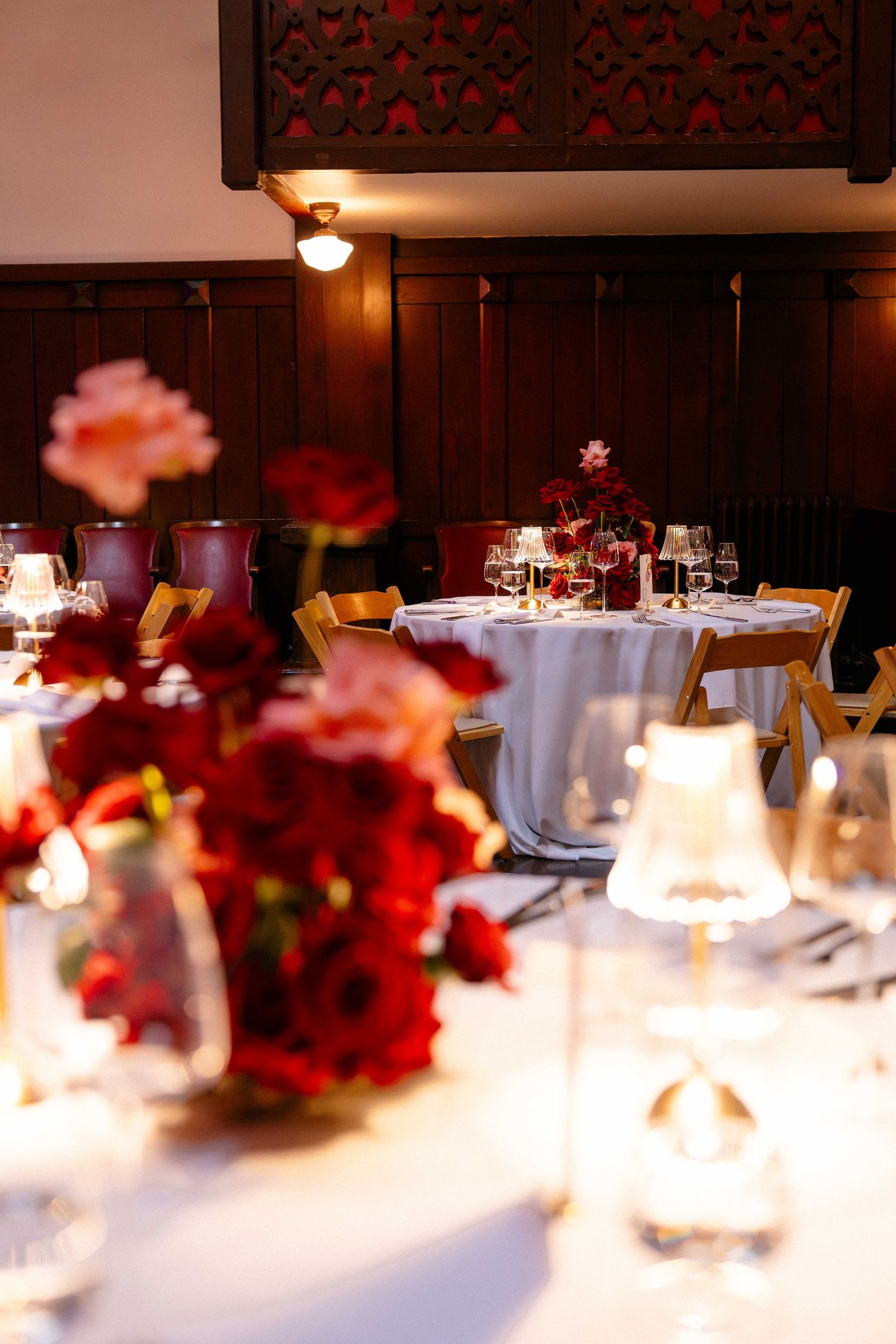Elegant wedding reception with red rose centerpieces, candlelight, and white tablecloths in a warmly lit wood-paneled venue.