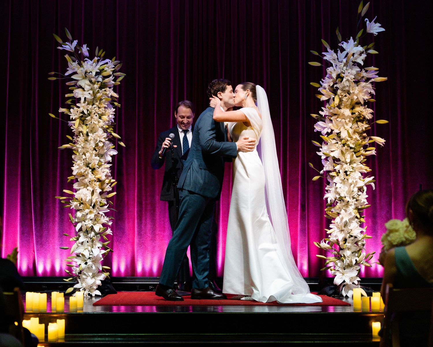 Bride and groom share first kiss at elegant wedding altar with floral columns and purple draped backdrop.