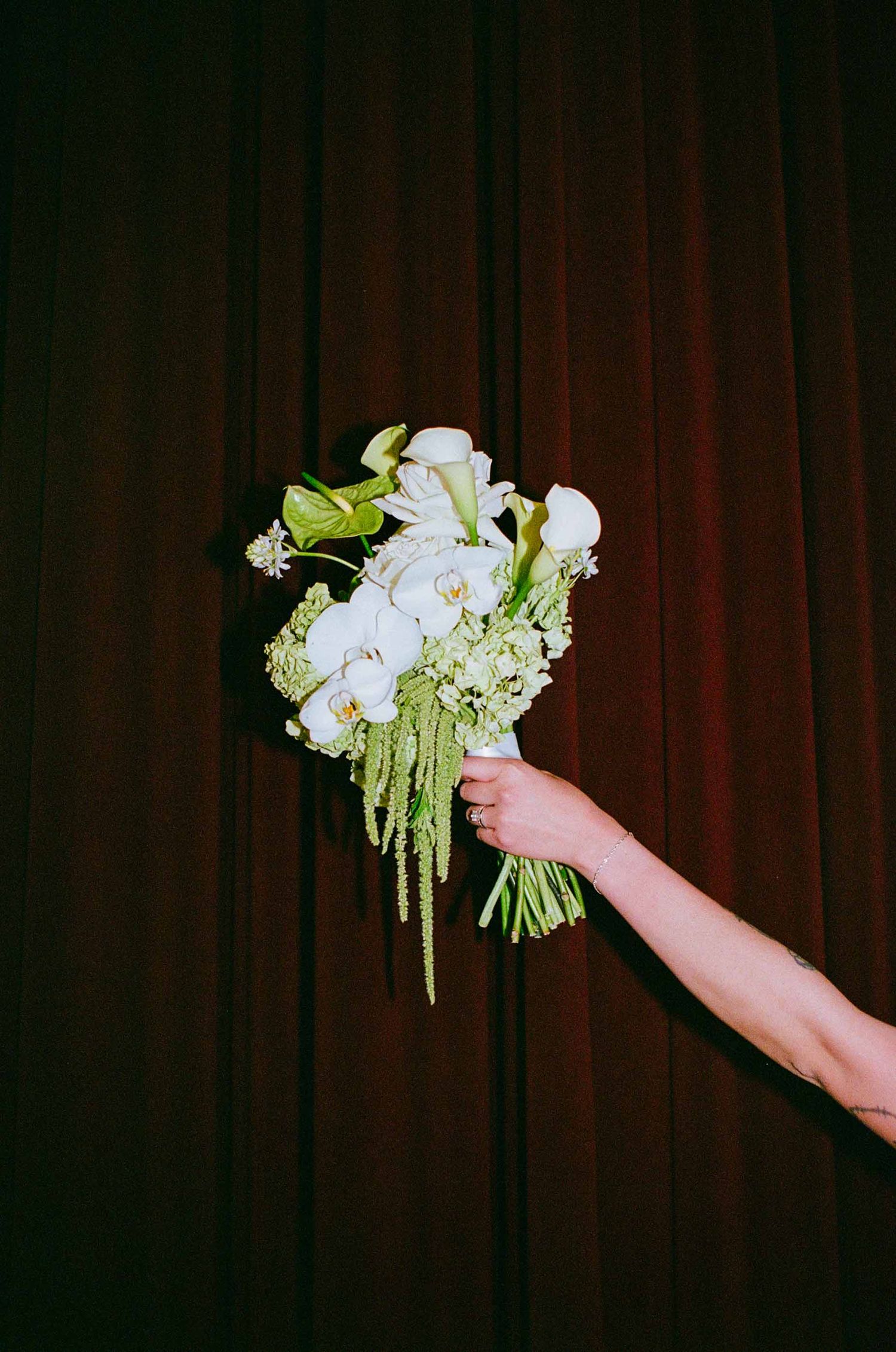 Hand holds elegant white orchid and green bouquet against dark red curtain backdrop.