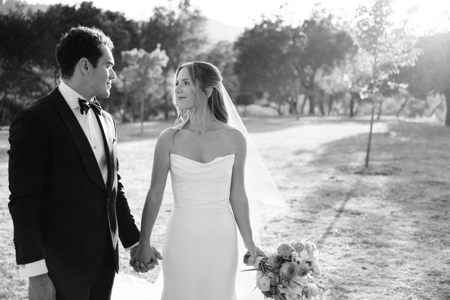 Black and white photo of bride and groom holding hands while walking through a sunlit outdoor venue.