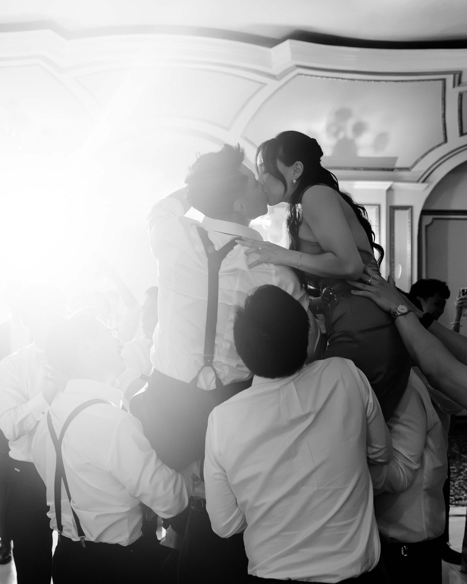 Black and white photo of a couple sharing a romantic kiss while being lifted by wedding guests in a bright venue.