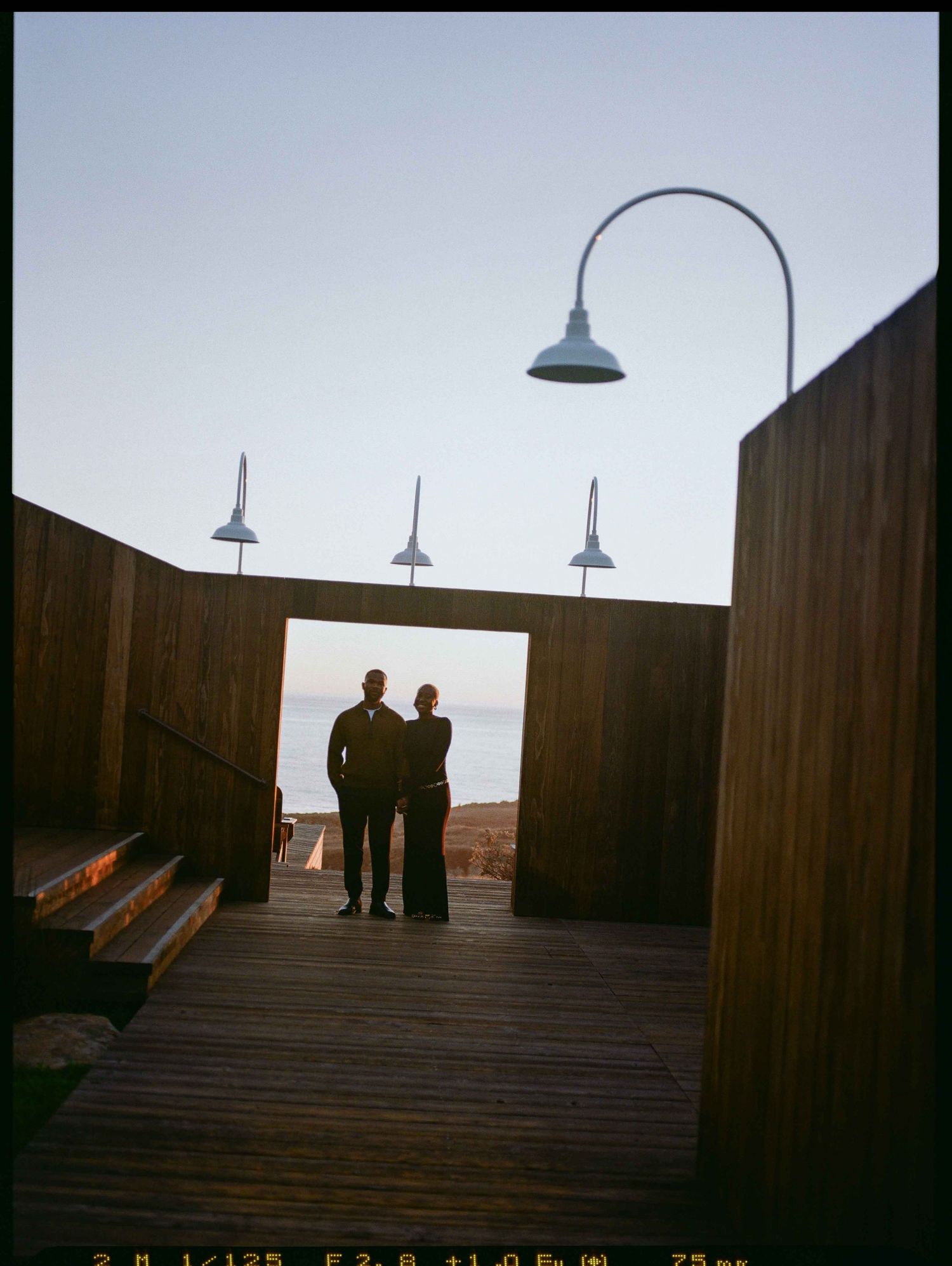 Couple silhouetted in wooden boardwalk archway at dusk with gooseneck lamps and ocean view backdrop.