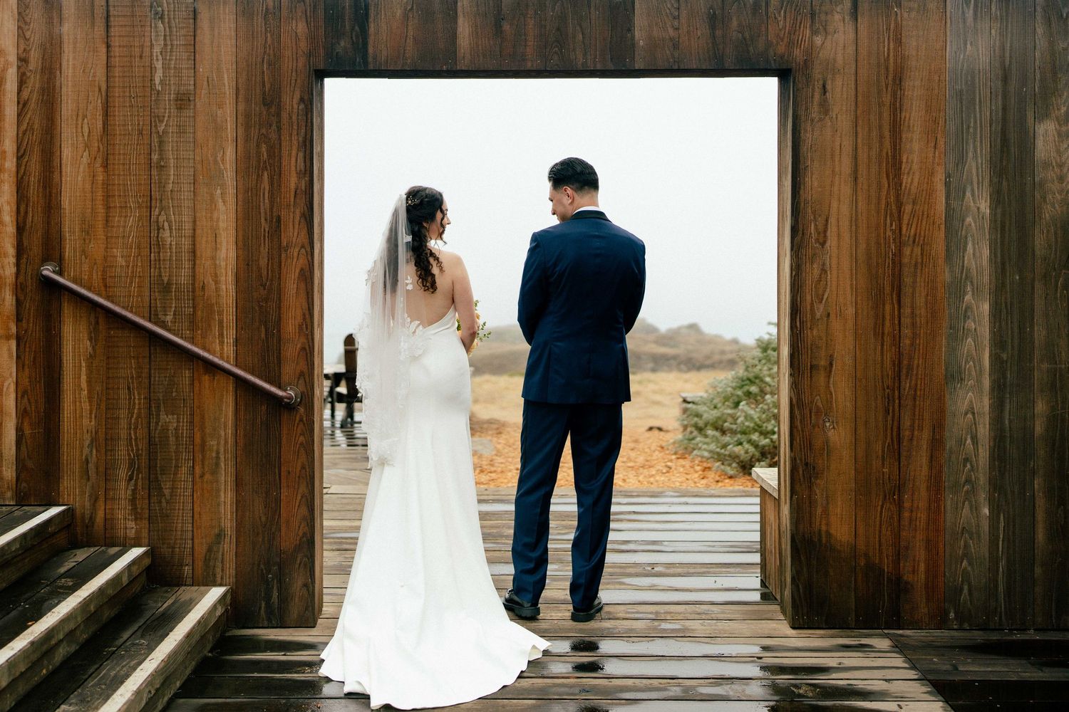 Bride and groom standing hand in hand in wooden doorway overlooking scenic outdoor landscape on wedding day.