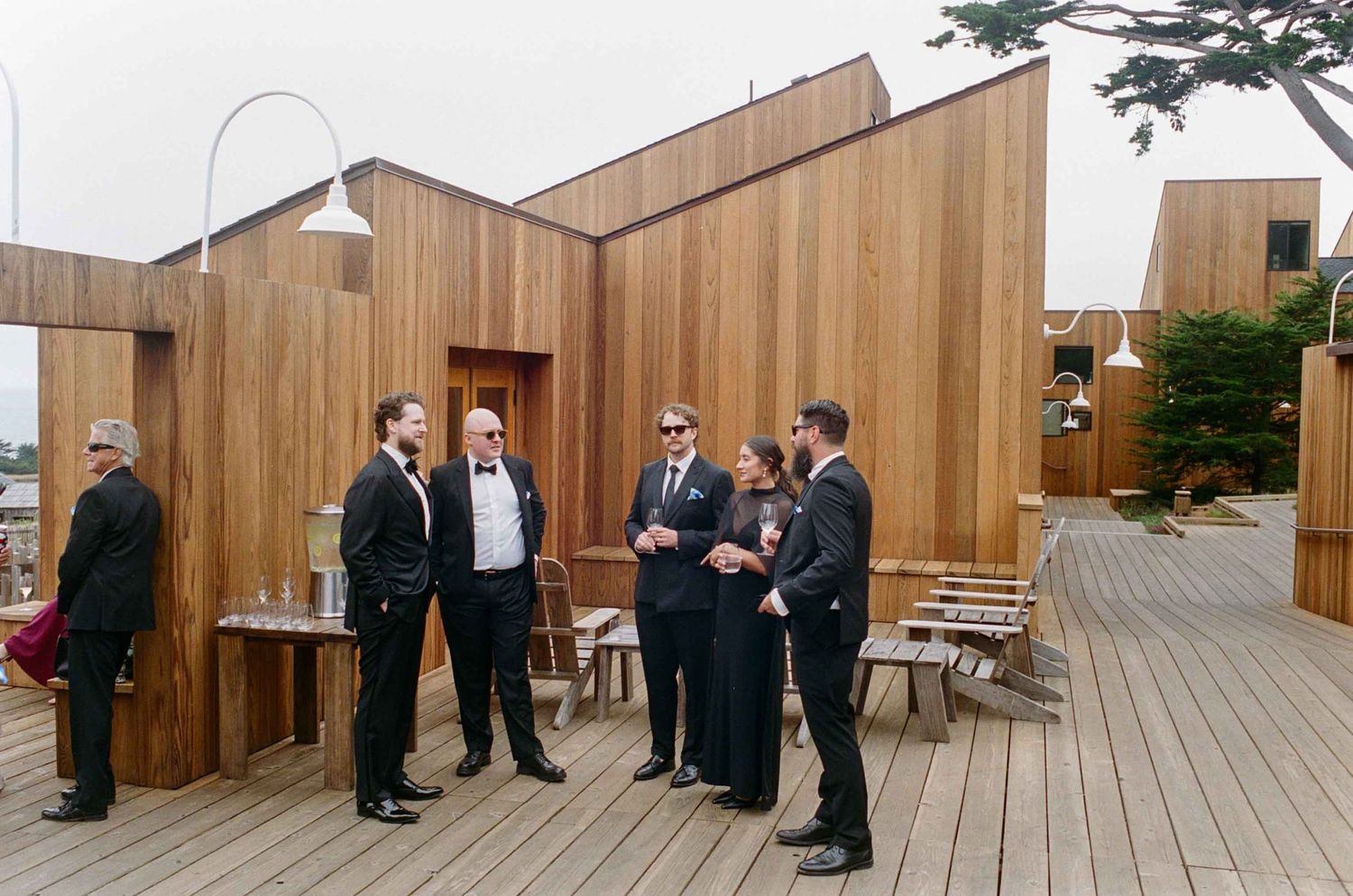 Groomsmen in black suits socializing on a wooden deck outside a modern timber building on a cloudy day.
