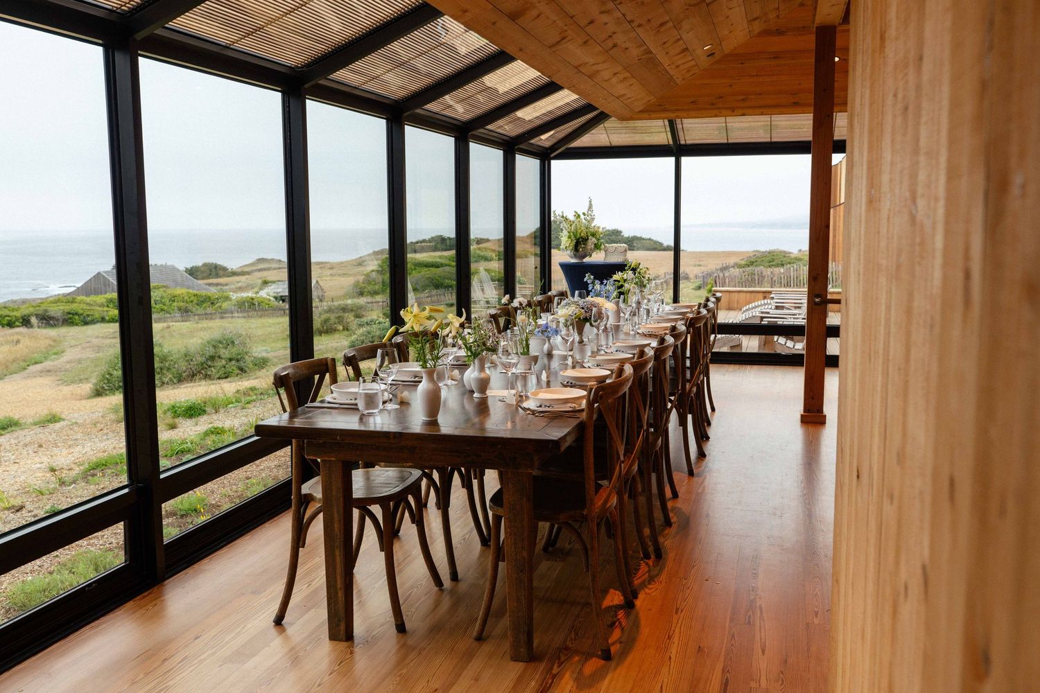 Elegant dining room with long wooden table, cross-back chairs, and panoramic ocean views through floor-to-ceiling windows.