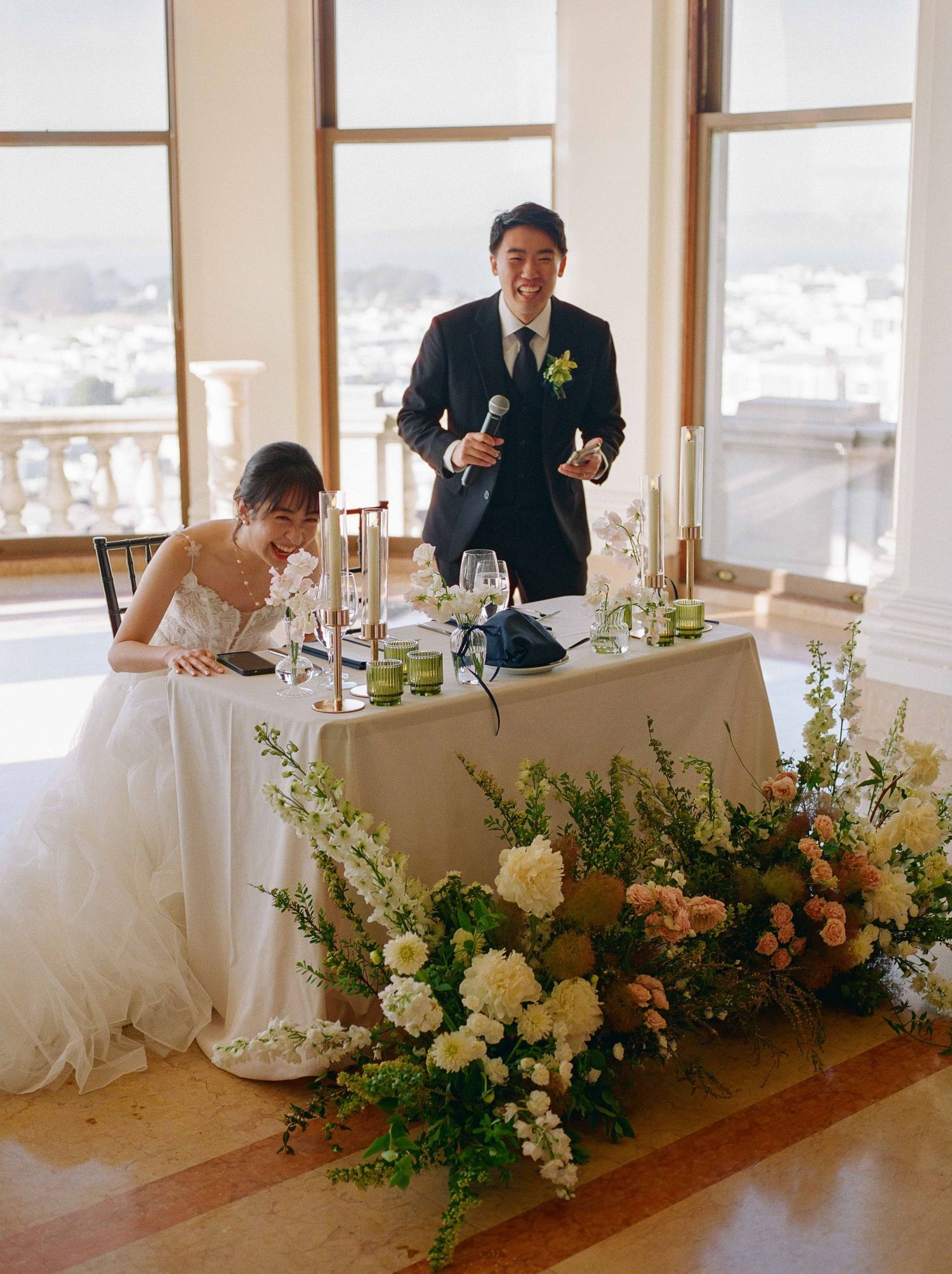 Bride and groom at floral-adorned sweetheart table, groom giving speech at elegant wedding reception.