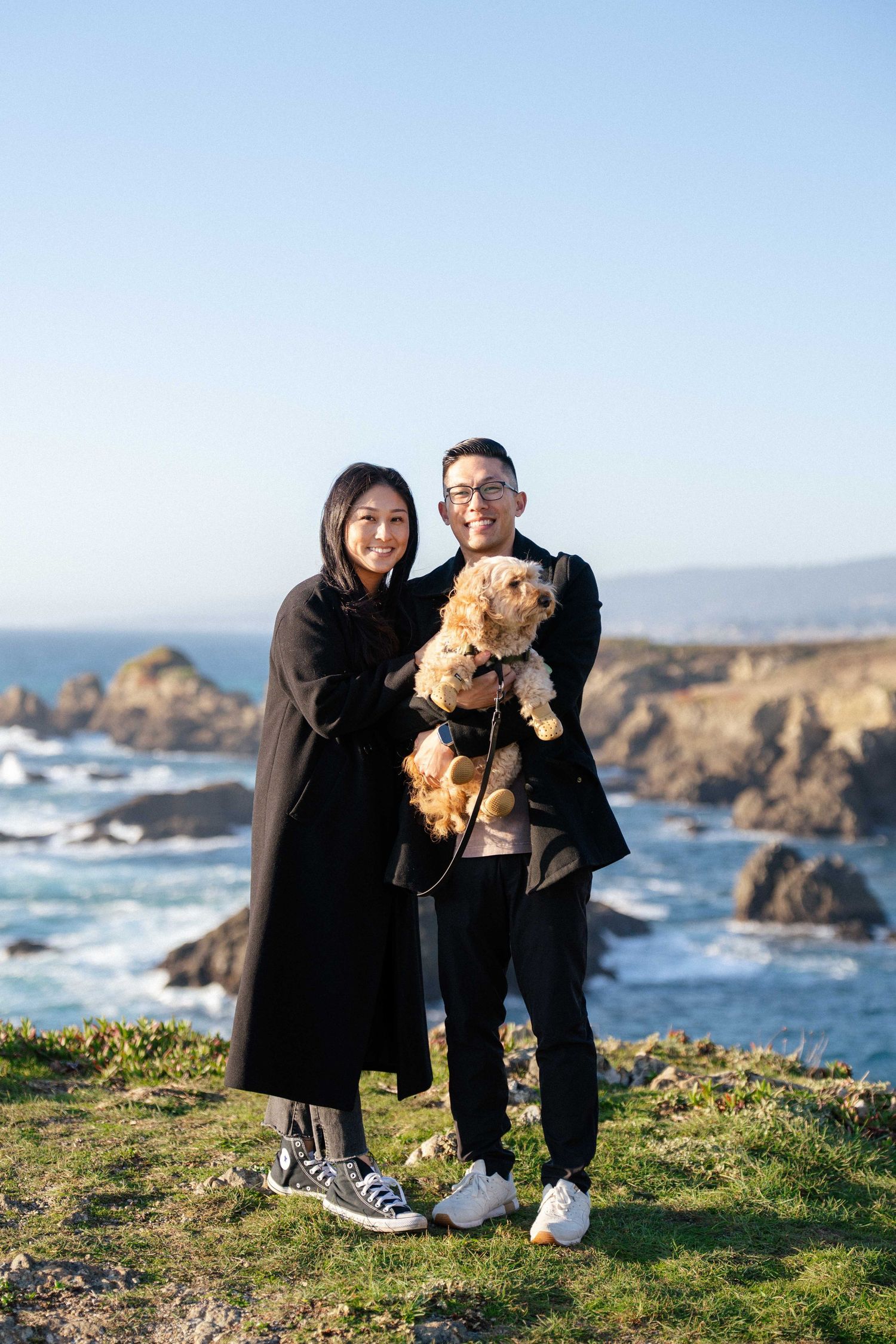 Couple in black outfits holding a fluffy dog on a coastal cliff with rocky ocean views and clear blue sky.