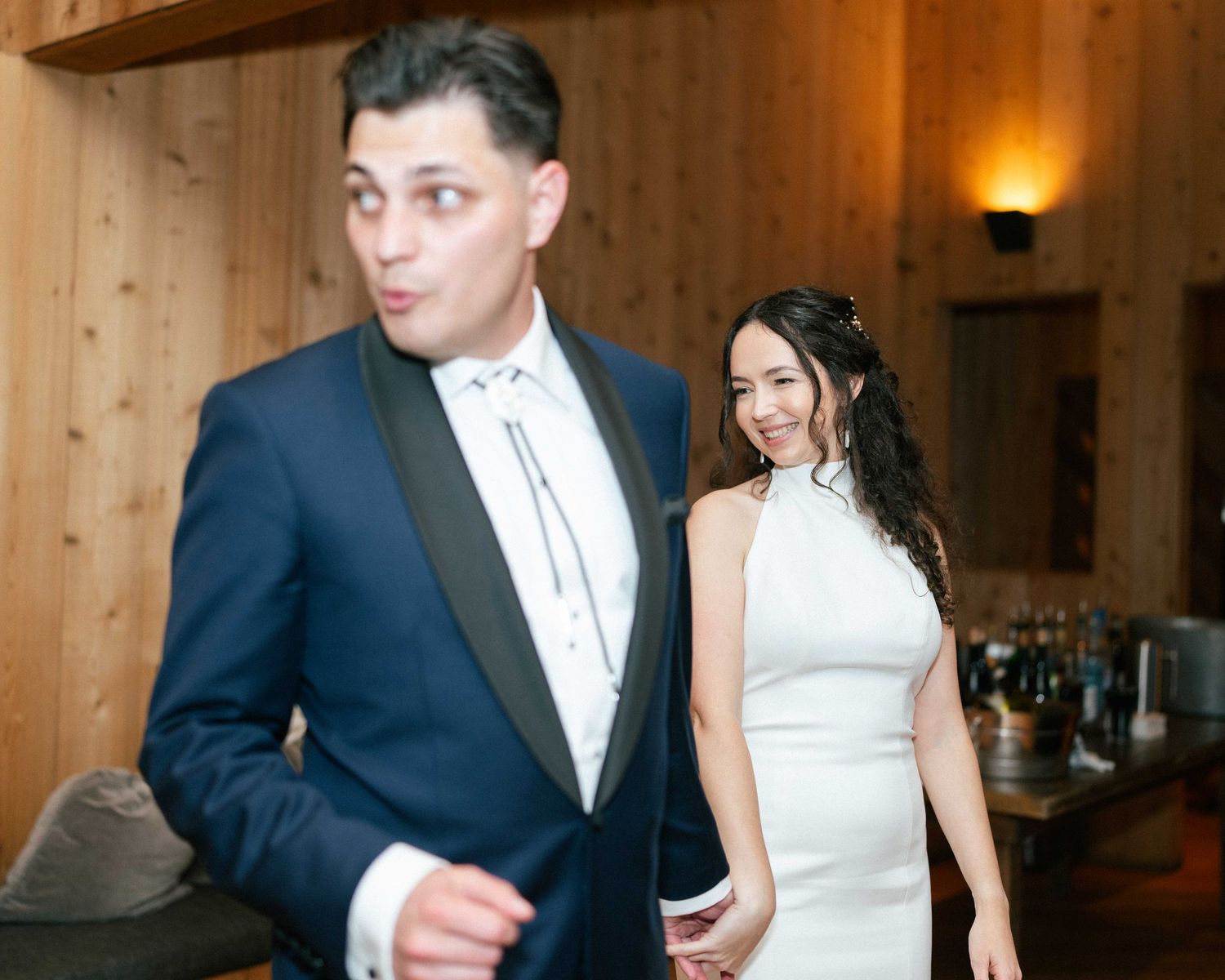 Bride in white halter gown and groom in navy tuxedo at elegant wood-paneled wedding reception venue.