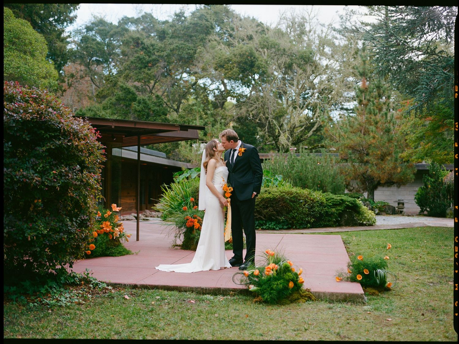 Bride and groom share a kiss on red terrace surrounded by orange flowers.