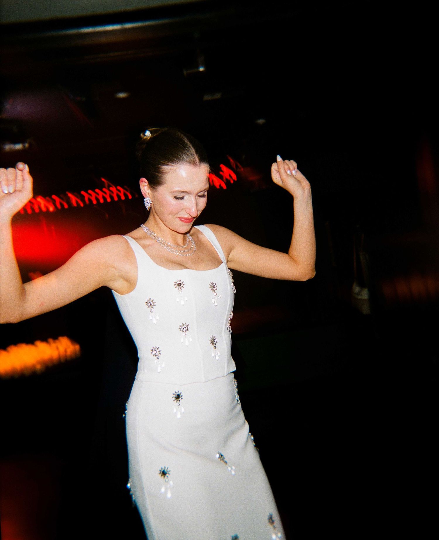 Woman in white embellished dress dancing joyfully at a dimly lit event with red ambient lighting.