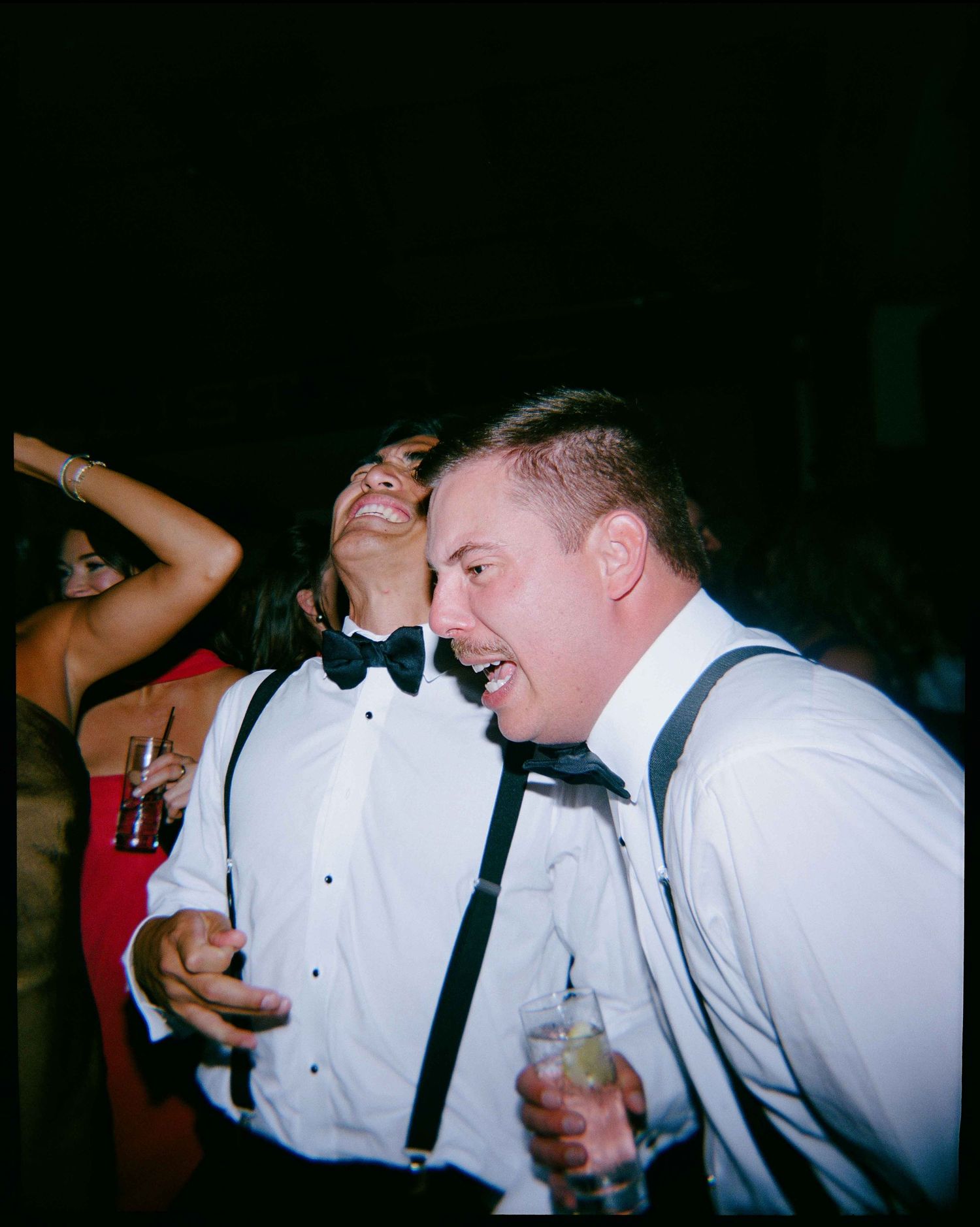 Two guests in bow ties and suspenders laughing and dancing at a formal wedding reception party.