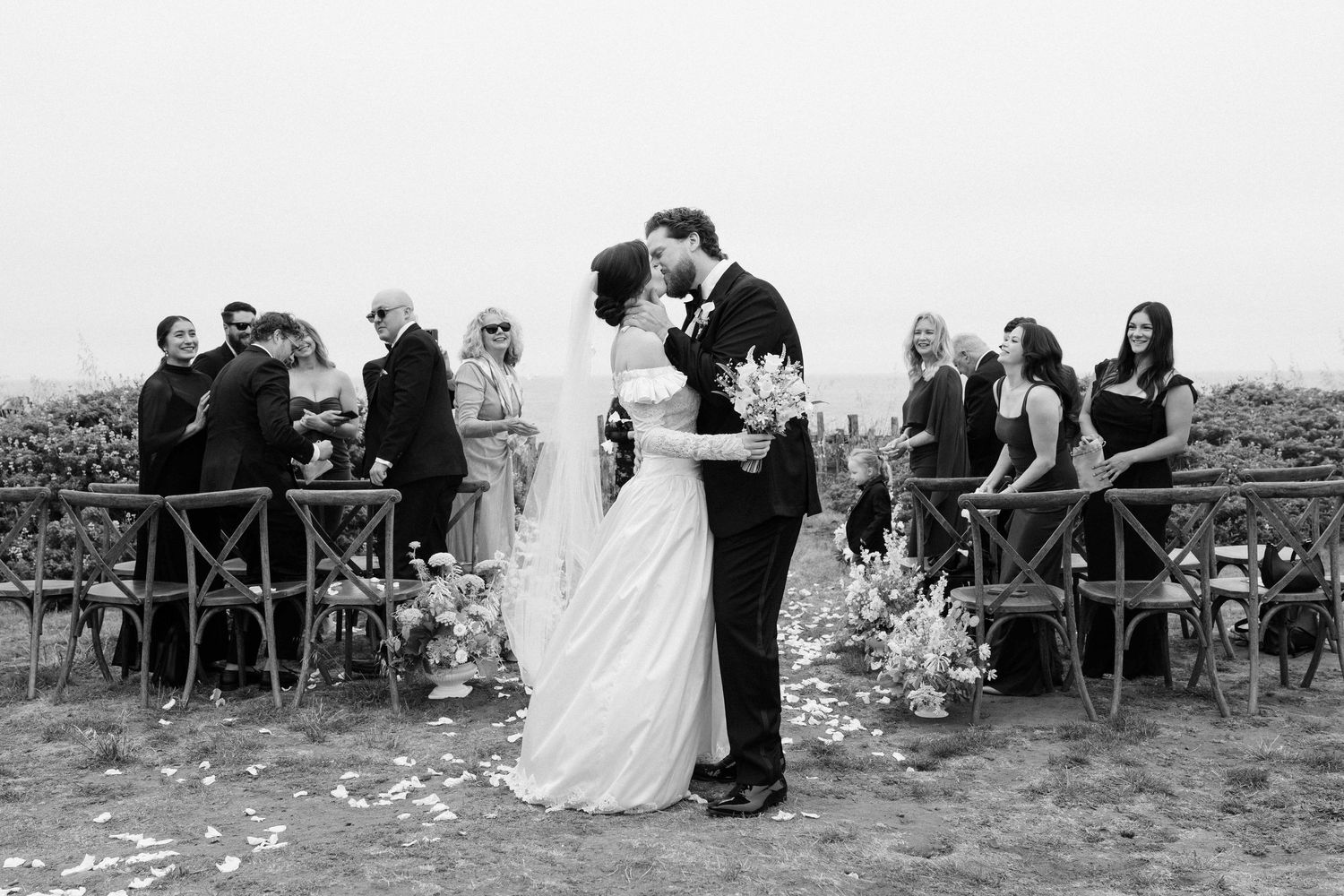 Black and white photo of bride and groom sharing first kiss at outdoor wedding ceremony with guests celebrating behind them.