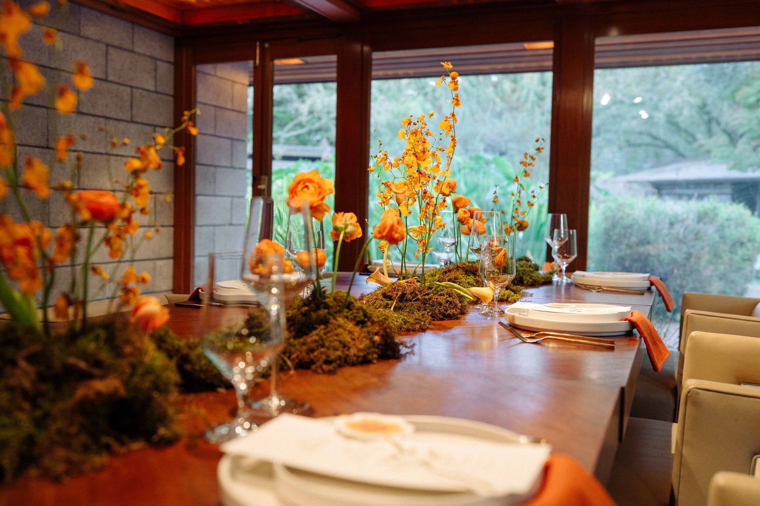 An elegant wedding reception table adorned with orange ranunculus and moss centerpieces beside large windows.