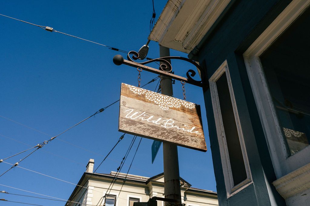 Vintage wooden business sign for WildBride hanging against bright blue sky with string lights.