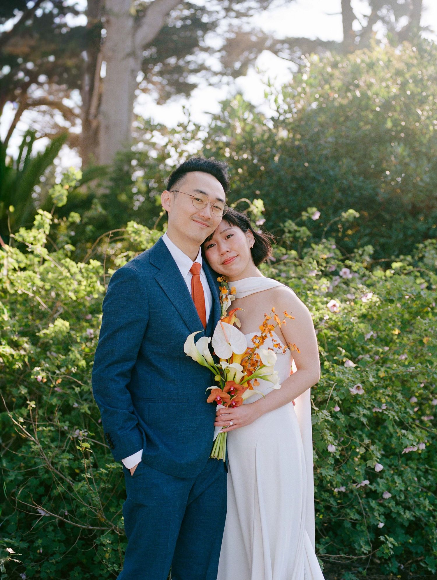 Couple poses in blue suit and white gown with orange bouquet amid lush greenery.