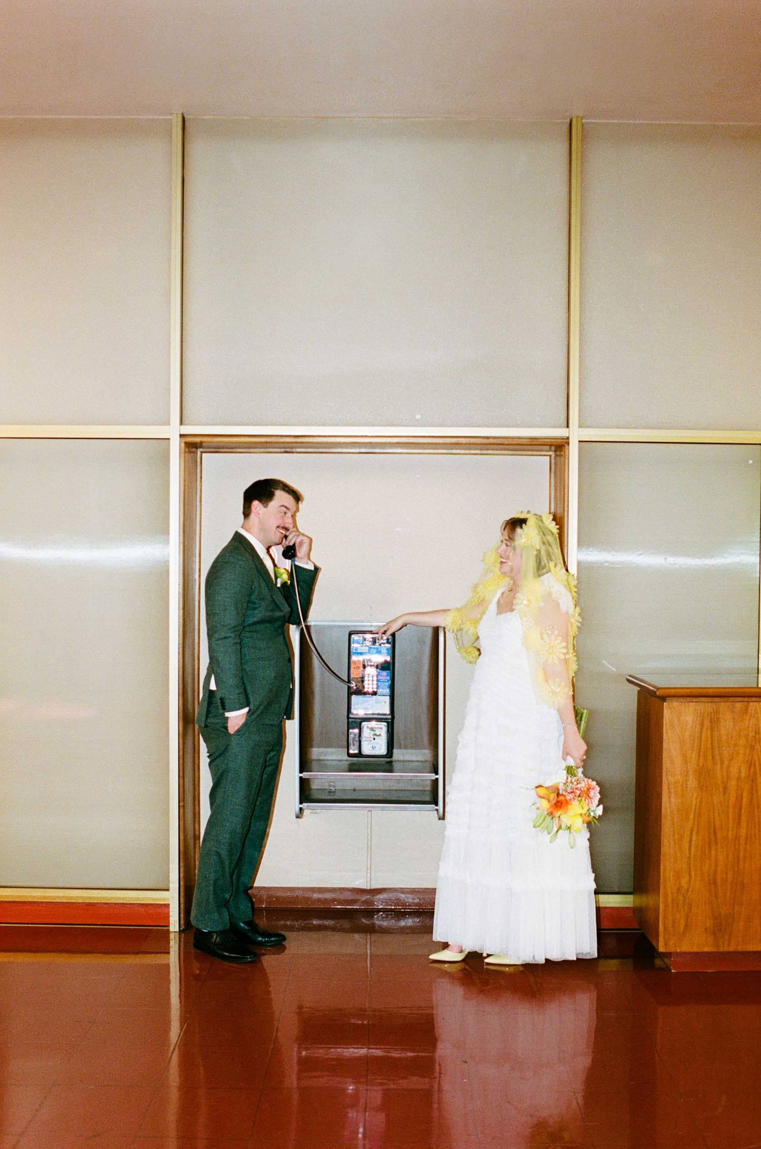 Bride in white gown and yellow veil holds flowers while groom in green suit uses a payphone at a wedding venue.