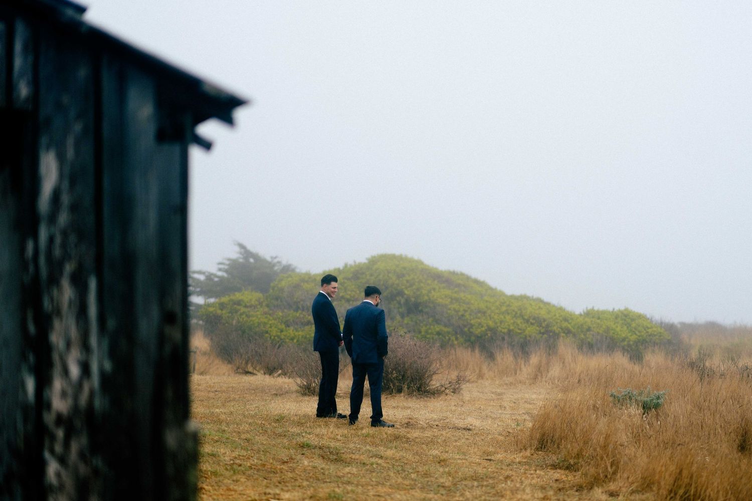 Two men in suits stand in a foggy coastal field near a weathered wooden structure.