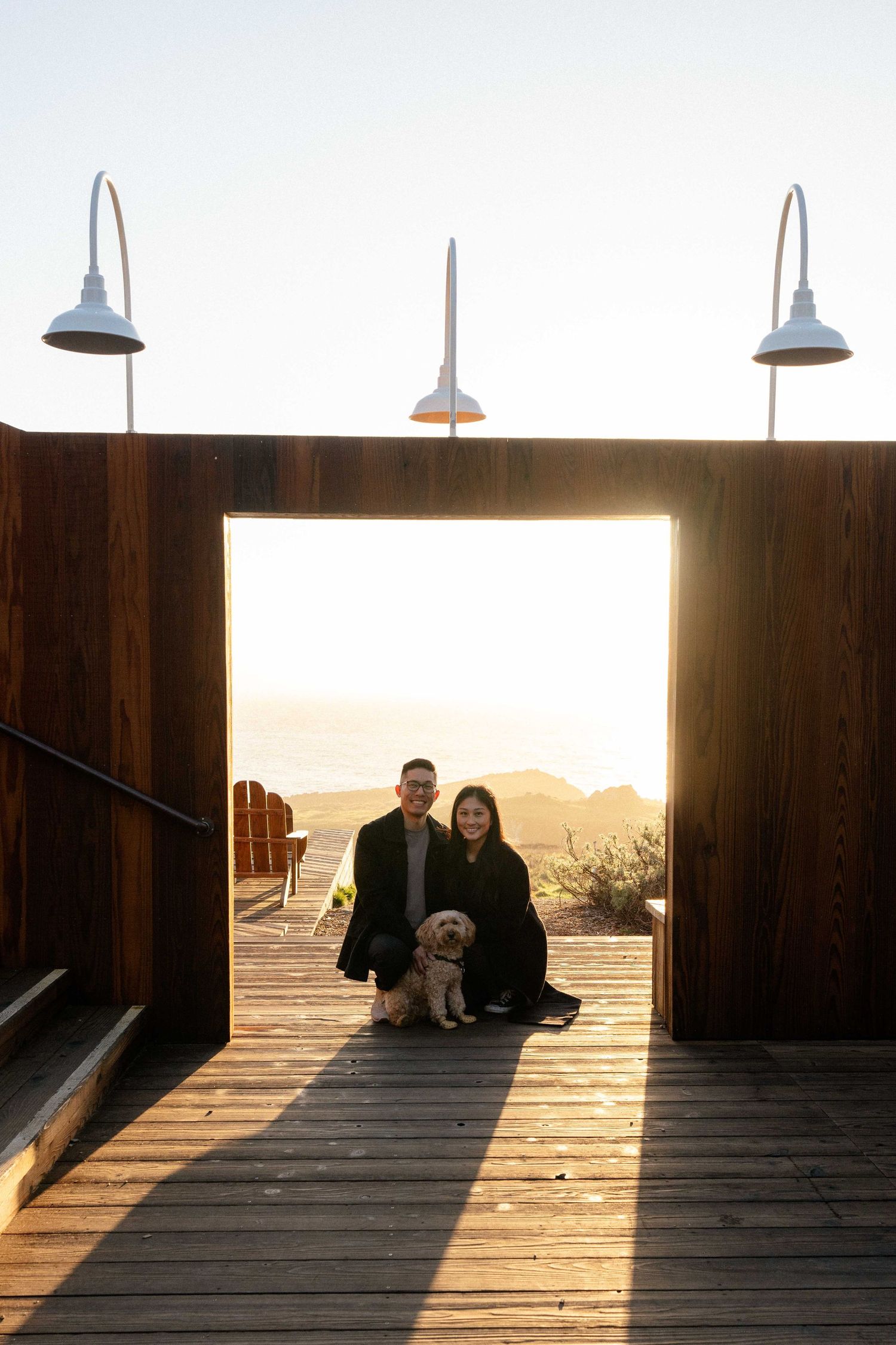 Couple and golden dog pose on wooden deck at sunset with ocean views through rustic doorway.