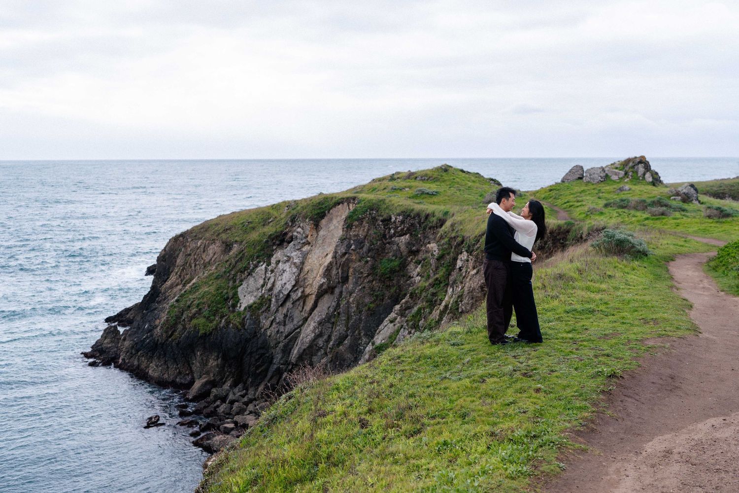 Couple embracing on a scenic coastal cliff with green grass, rocky shoreline, and calm ocean views under an overcast sky.