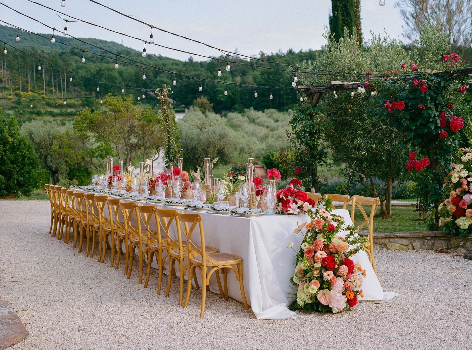 Elegant outdoor wedding reception with long table, wooden chairs, colorful floral arrangements, string lights, and lush greenery backdrop.
