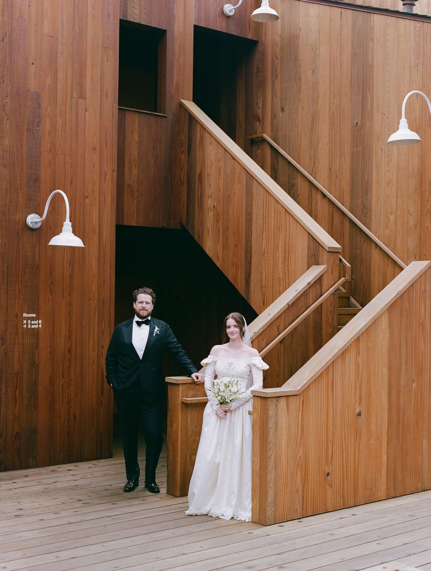 Bride and groom holding hands beside wooden staircase at rustic barn wedding venue.