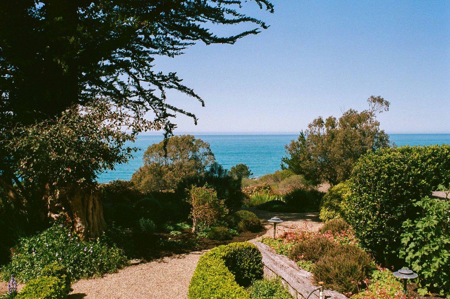 A scenic coastal garden path leads to the Mediterranean Sea with lush greenery and blue waters under clear skies.