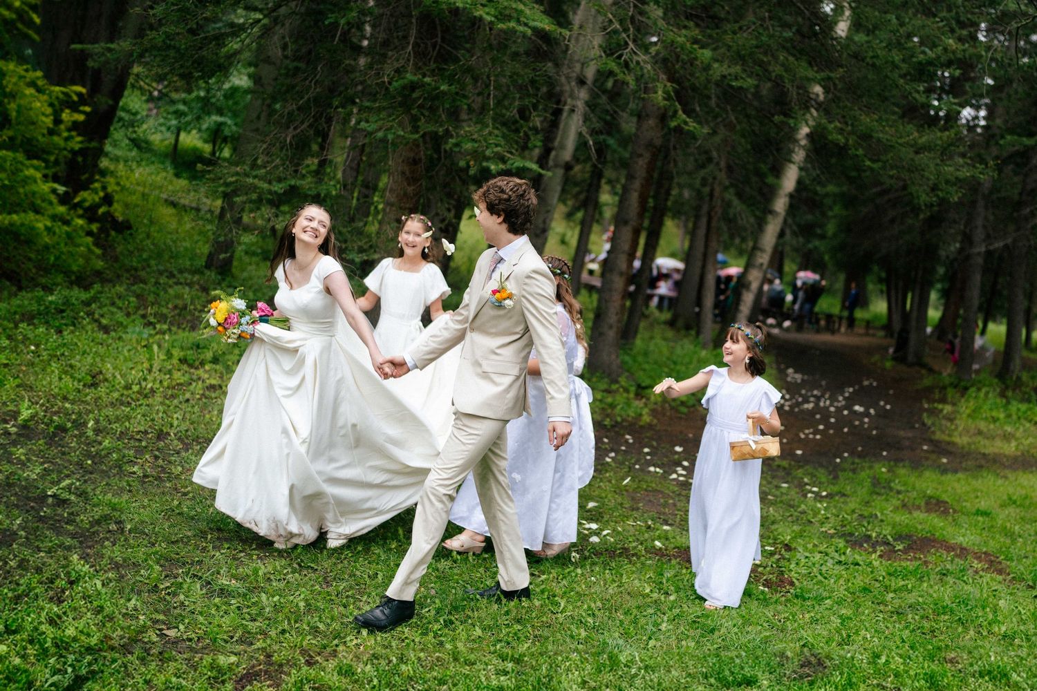 People in white outfits dance playfully in a lush green forest setting during a summer celebration.