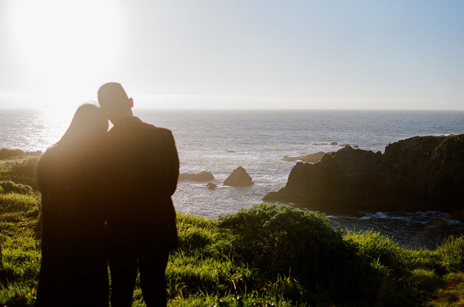 Couple silhouetted against golden sunset overlooking rocky Pacific coastline with lush green cliffs.