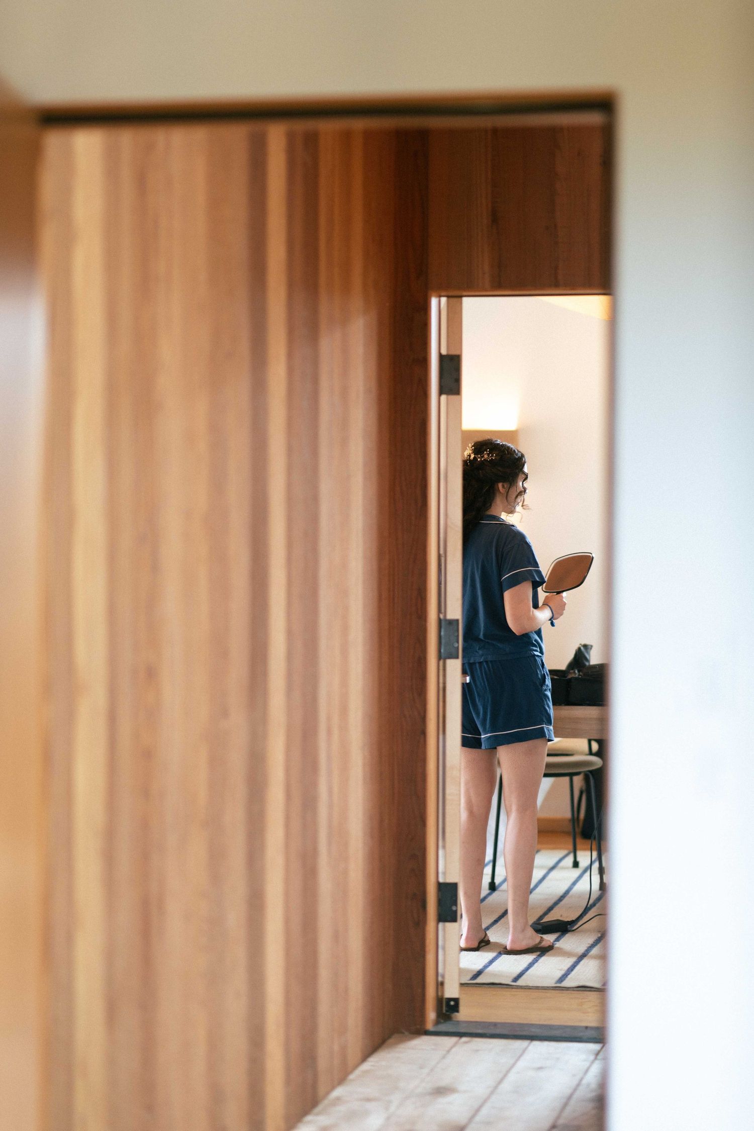 Woman in navy shorts holding a mirror, viewed through a wooden sliding door in a modern room.