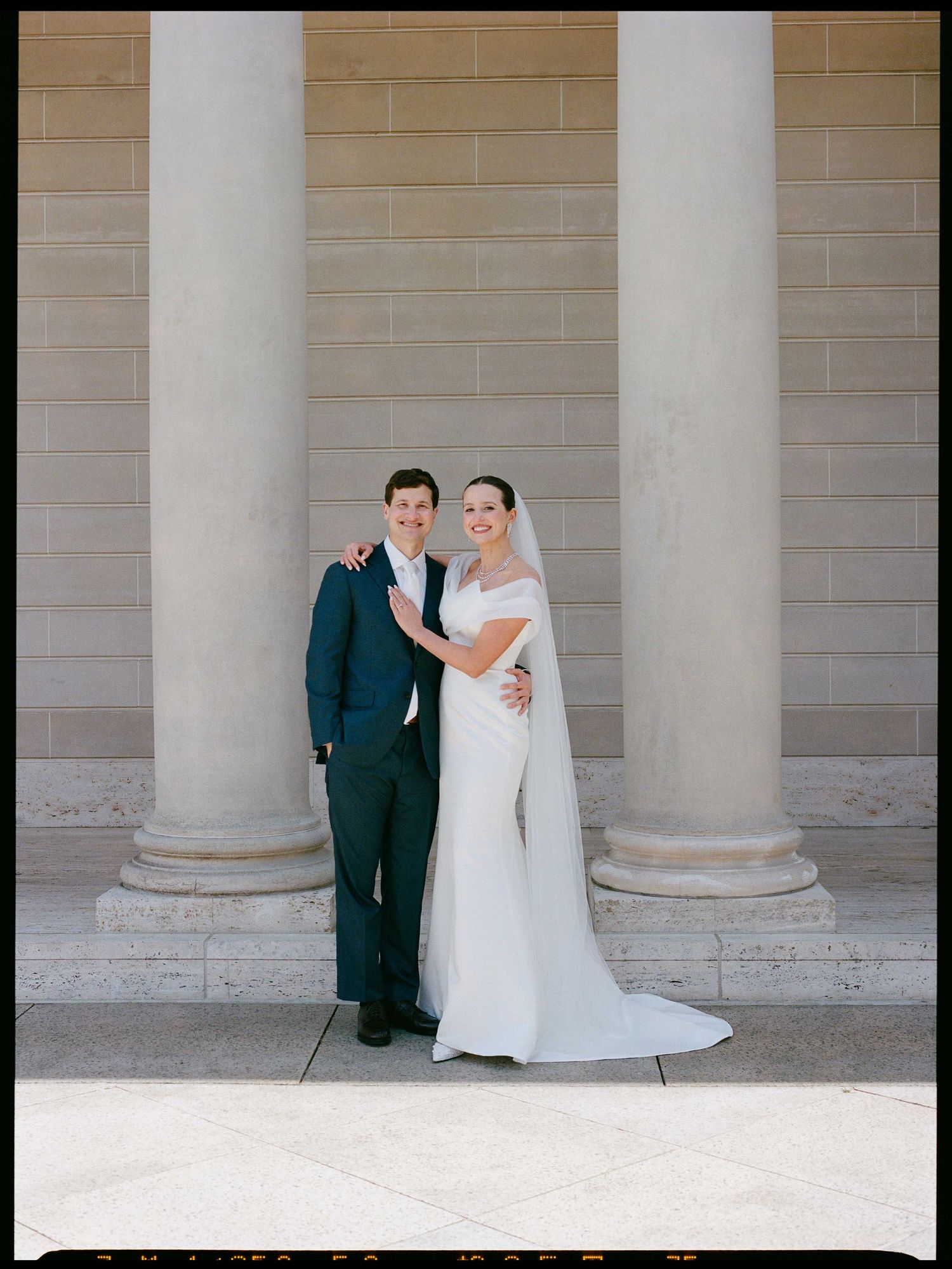 Bride in white gown and groom in navy suit pose together before grand stone columns in classic wedding portrait.