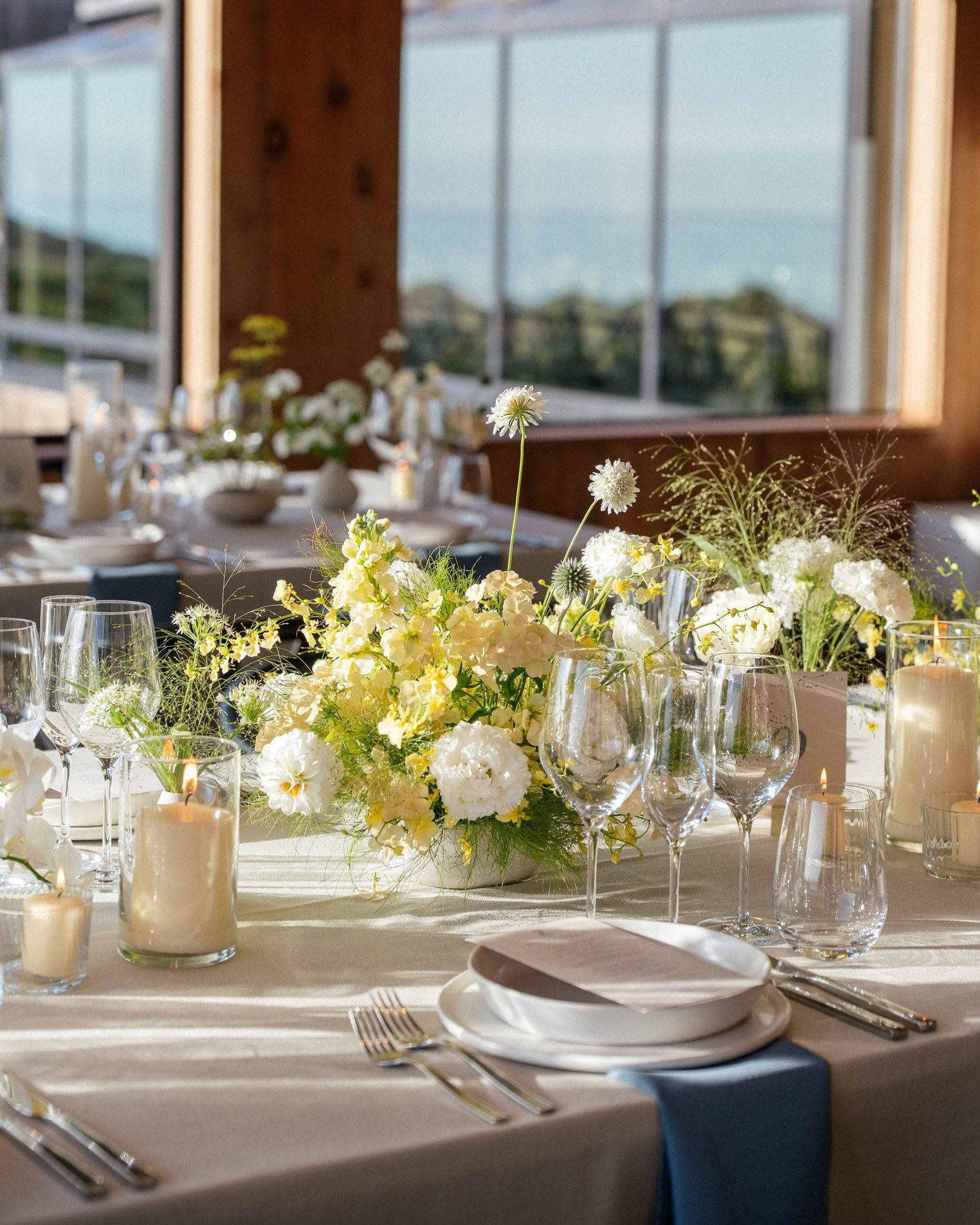 Elegant wedding reception table with white floral centerpiece, crystal glassware, candles, and blue napkins in a bright venue.