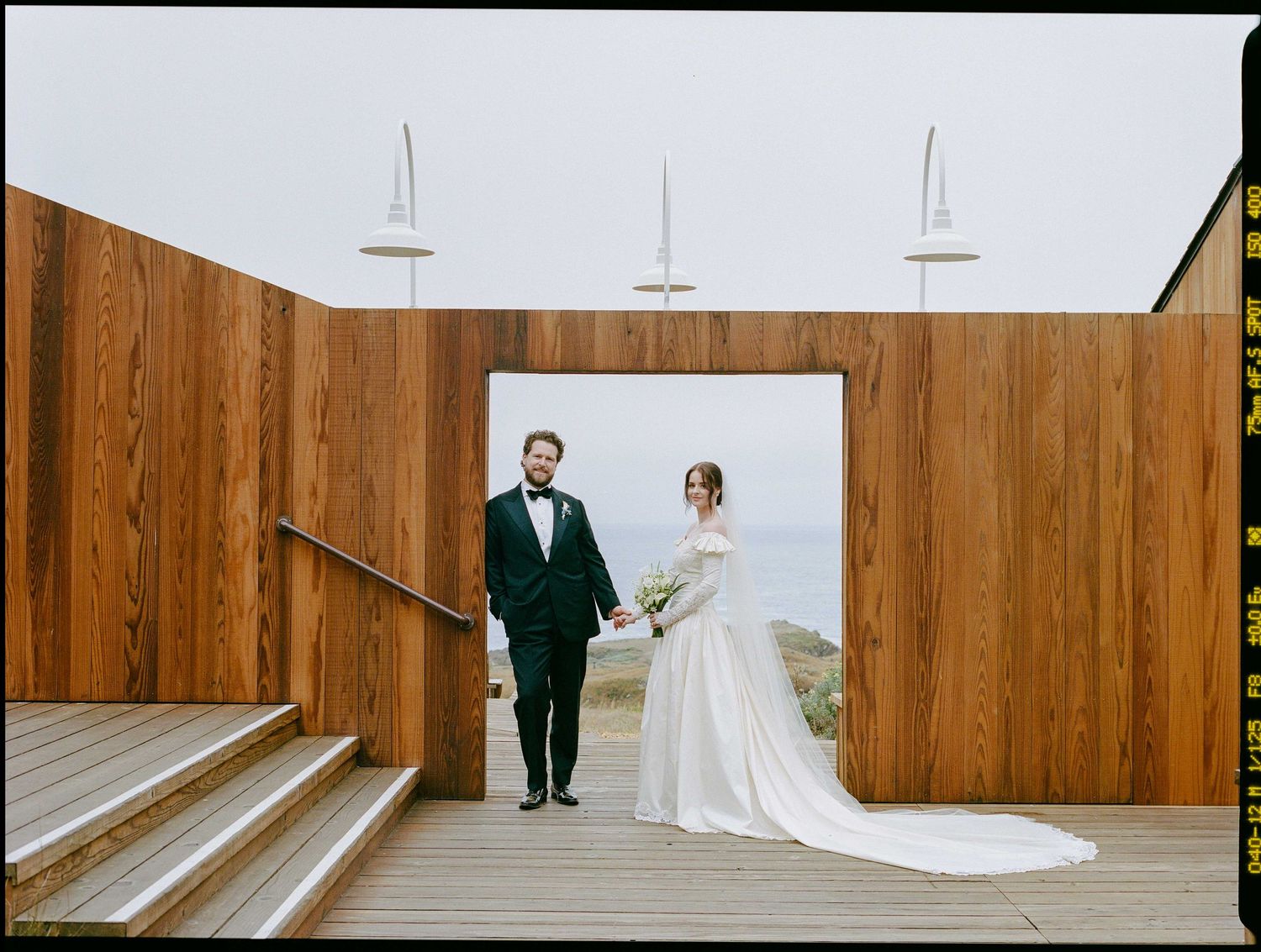 Bride and groom holding hands in a wooden chapel doorway with ocean views on their wedding day.