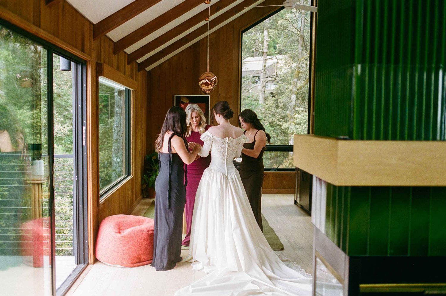 Bridesmaids helping bride into wedding gown in a modern wooden cabin with floor-to-ceiling windows.
