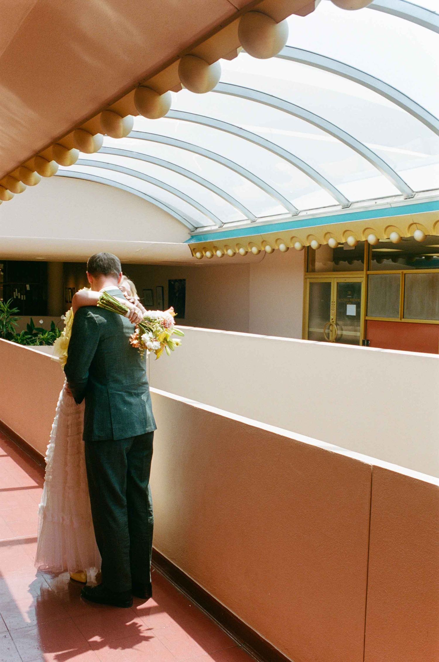 A couple embraces on a balcony, the bride holding flowers, under a curved glass ceiling in a elegant venue.