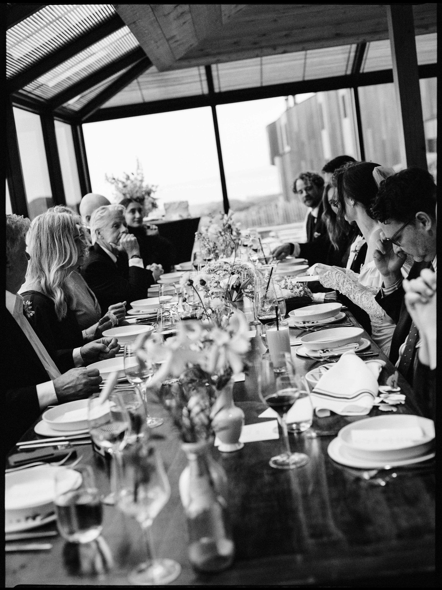 Black and white photo of elegant dinner party guests seated at a long decorated table in a glass conservatory.