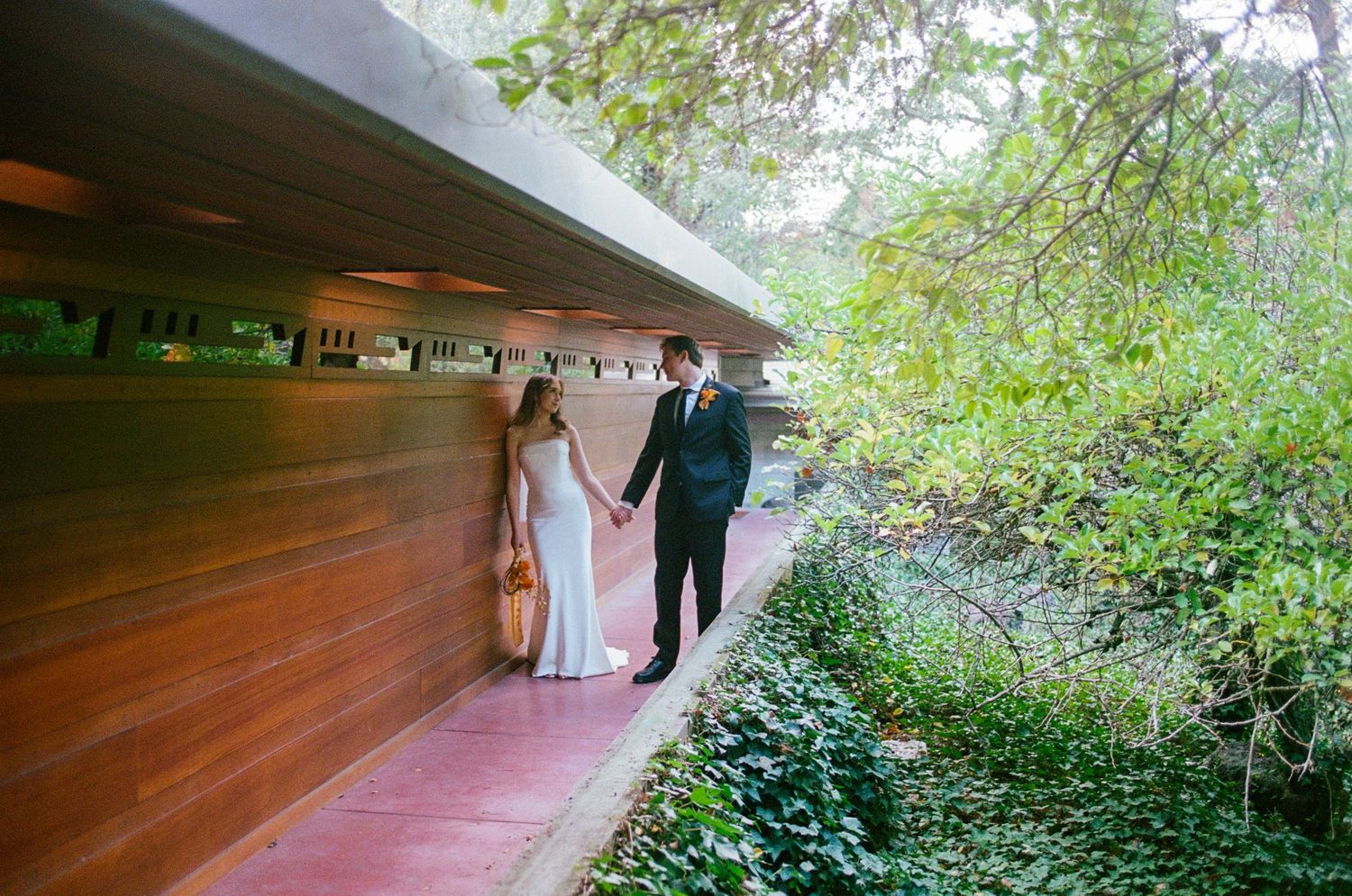 Bride and groom hold hands while walking along a red path beside a wooden overhang surrounded by greenery.