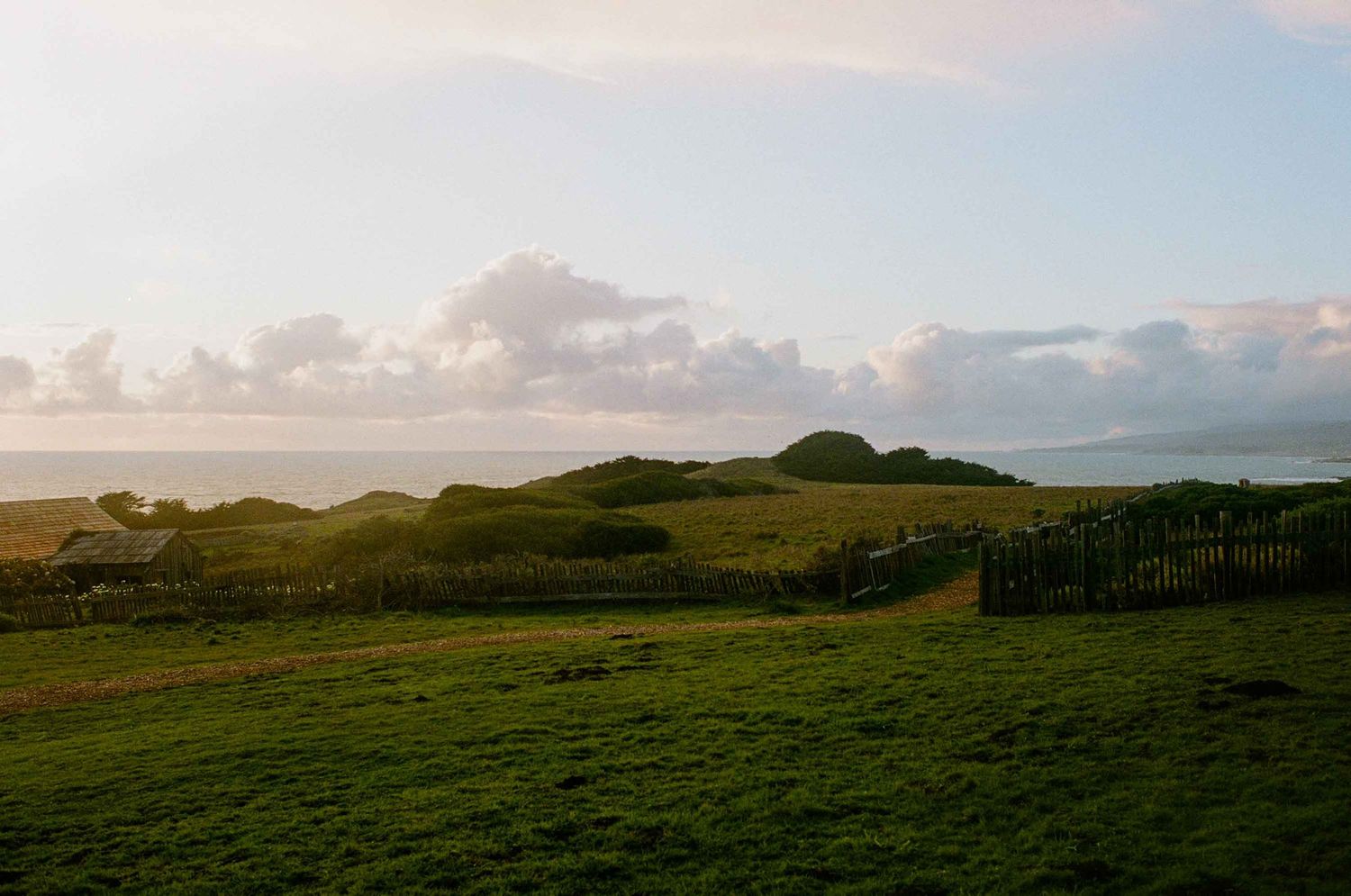 Golden hour light illuminates rolling green hills and a wooden fence near a coastal landscape.