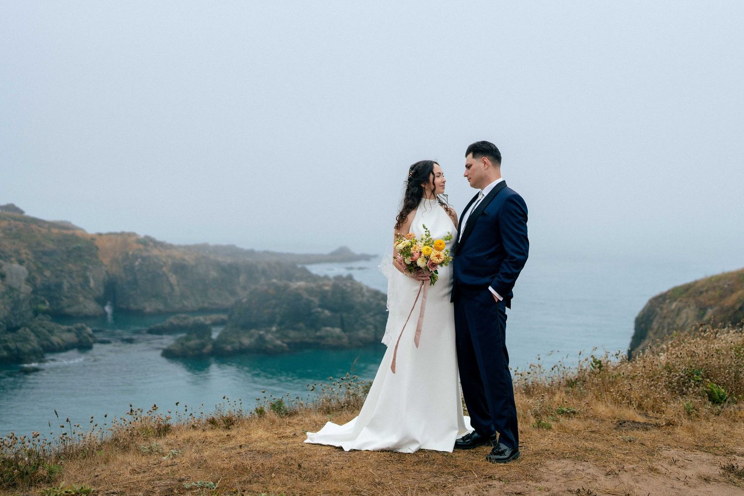 A bride and groom stand together on a dramatic coastal cliff overlooking misty blue ocean waters.