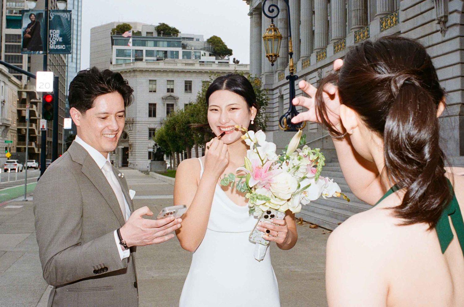 Bride applying lipstick outside of City Hall while her sister looks at her and the groom looks at his phone.