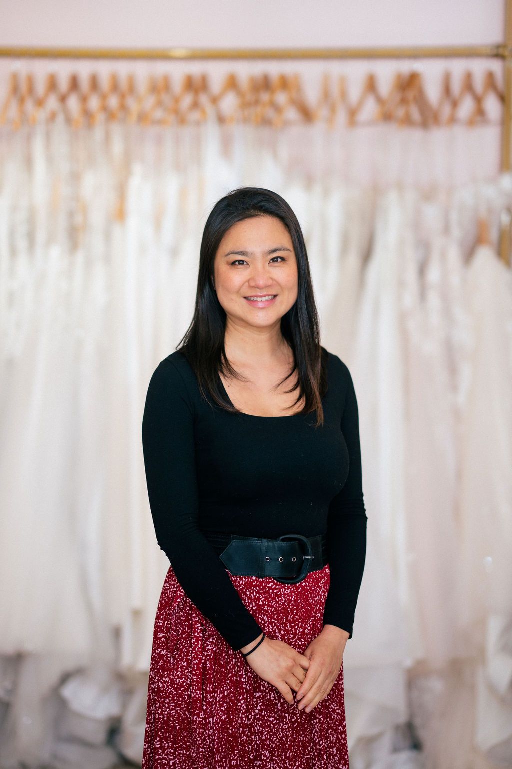 A smiling person in a black top and red sequin skirt poses in front of wedding dresses.