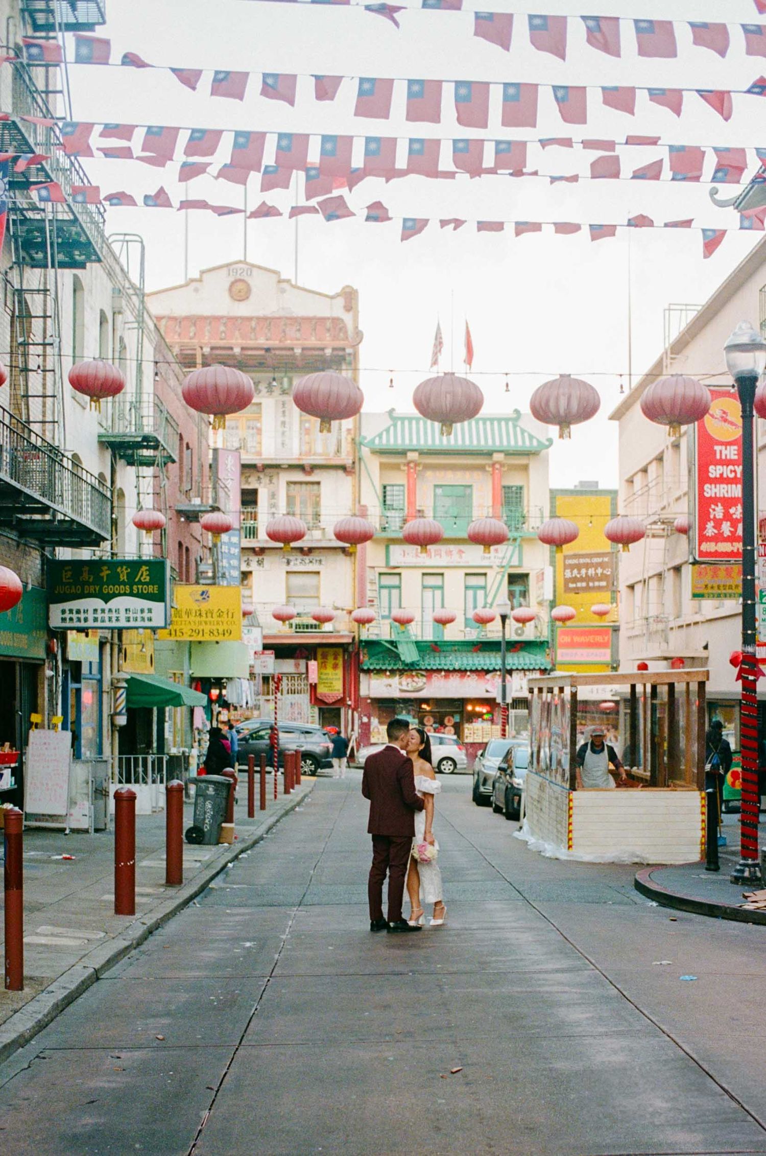 Bride and groom kissing in Chinatown with traditional architecture and red lanterns overhead.